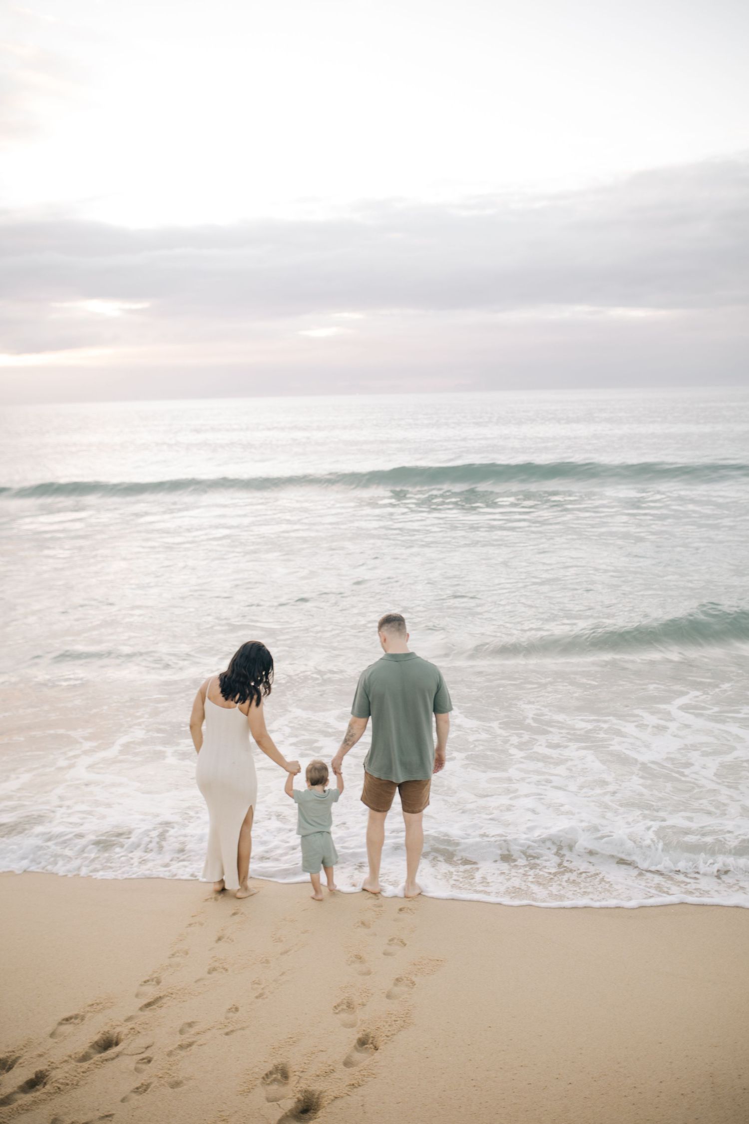 A family of three stands at the ocean's edge watching waves roll in at sunset.