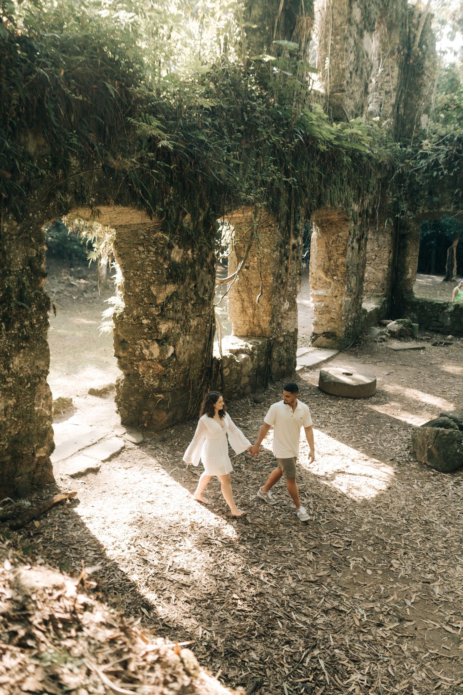 A couple walks hand in hand through ancient stone arches covered in vines.