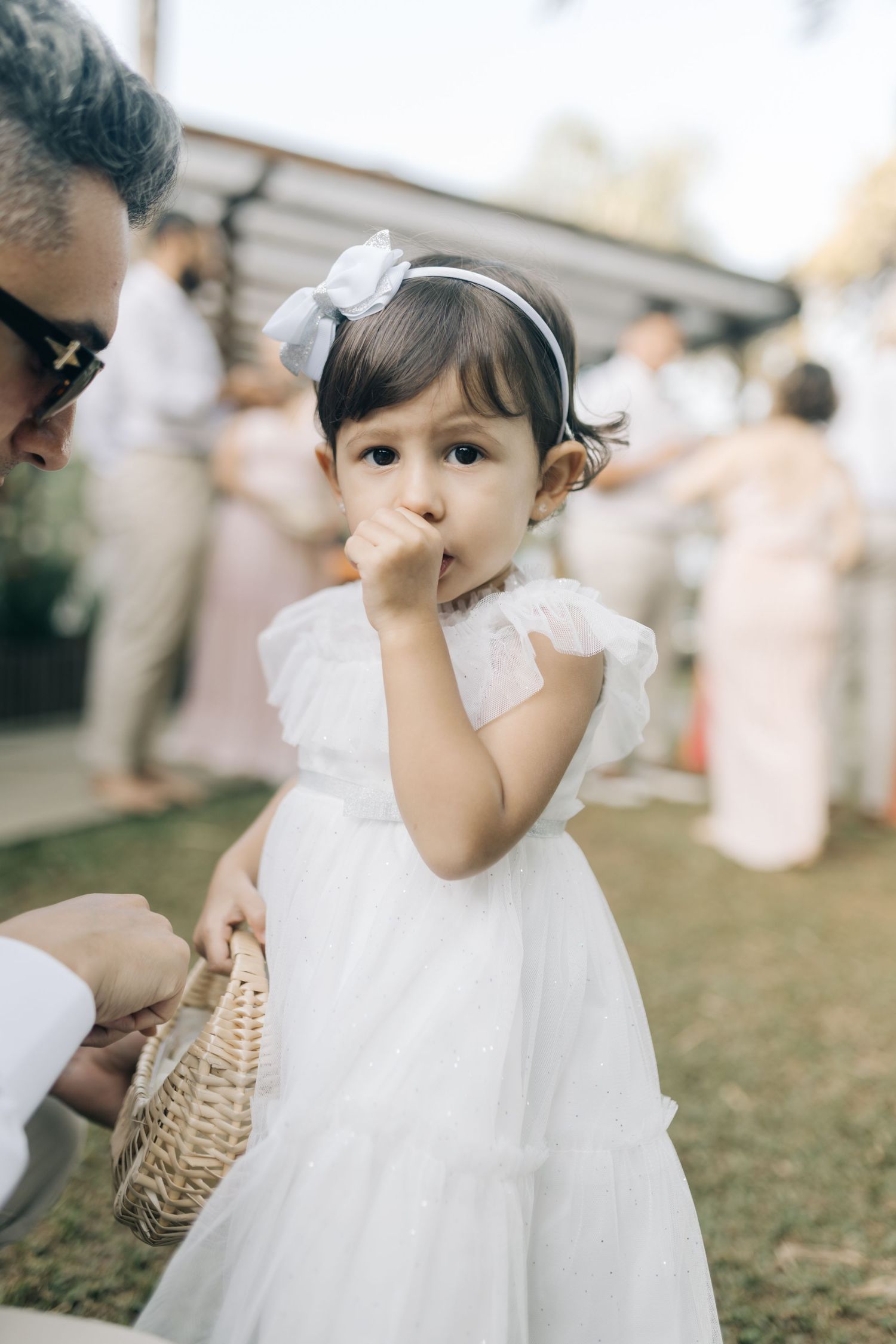A small child in white dress holds a wicker basket at an outdoor celebration.