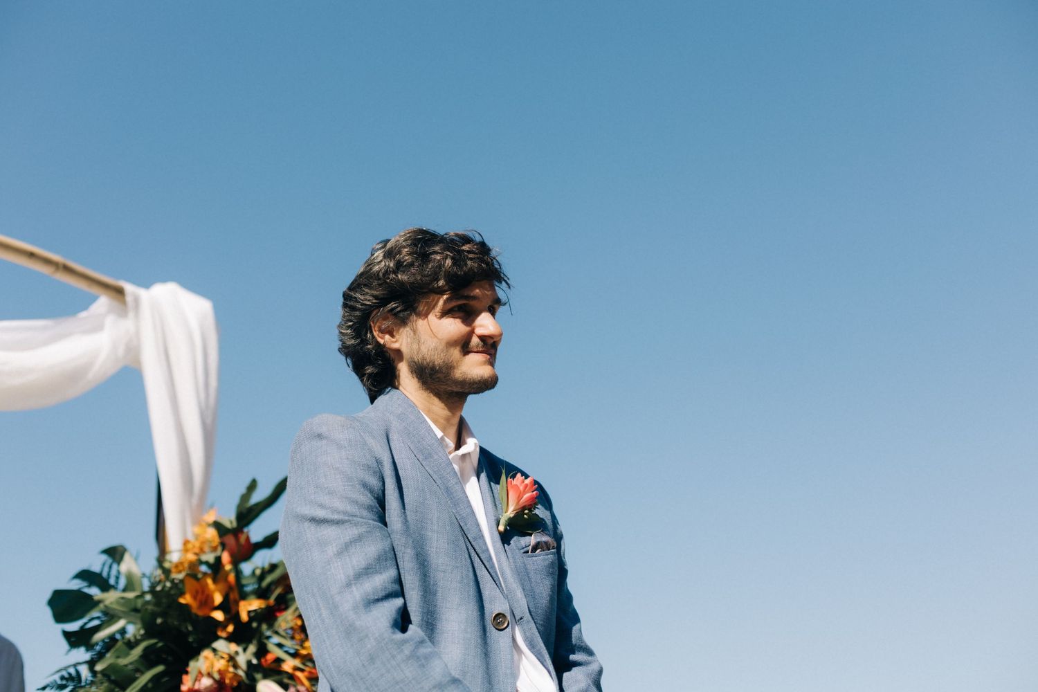 Person in a light blue suit stands against a bright blue sky at an outdoor wedding.