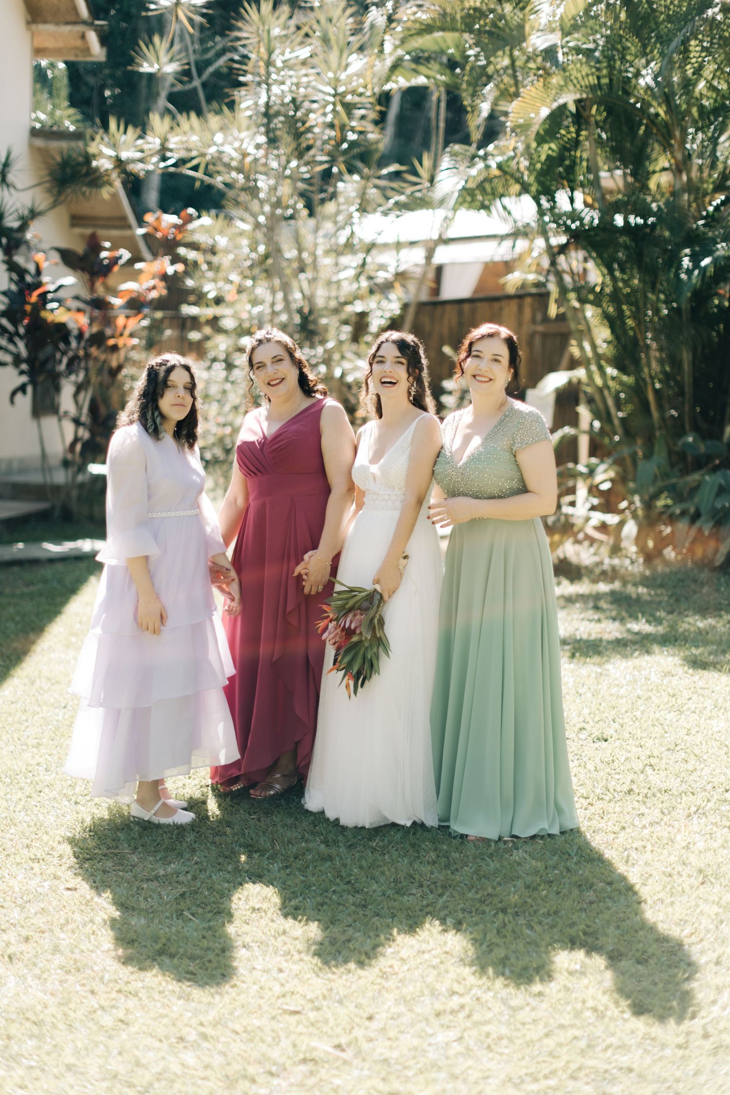 Wedding party in pastel and burgundy dresses posing together on a sunny lawn.