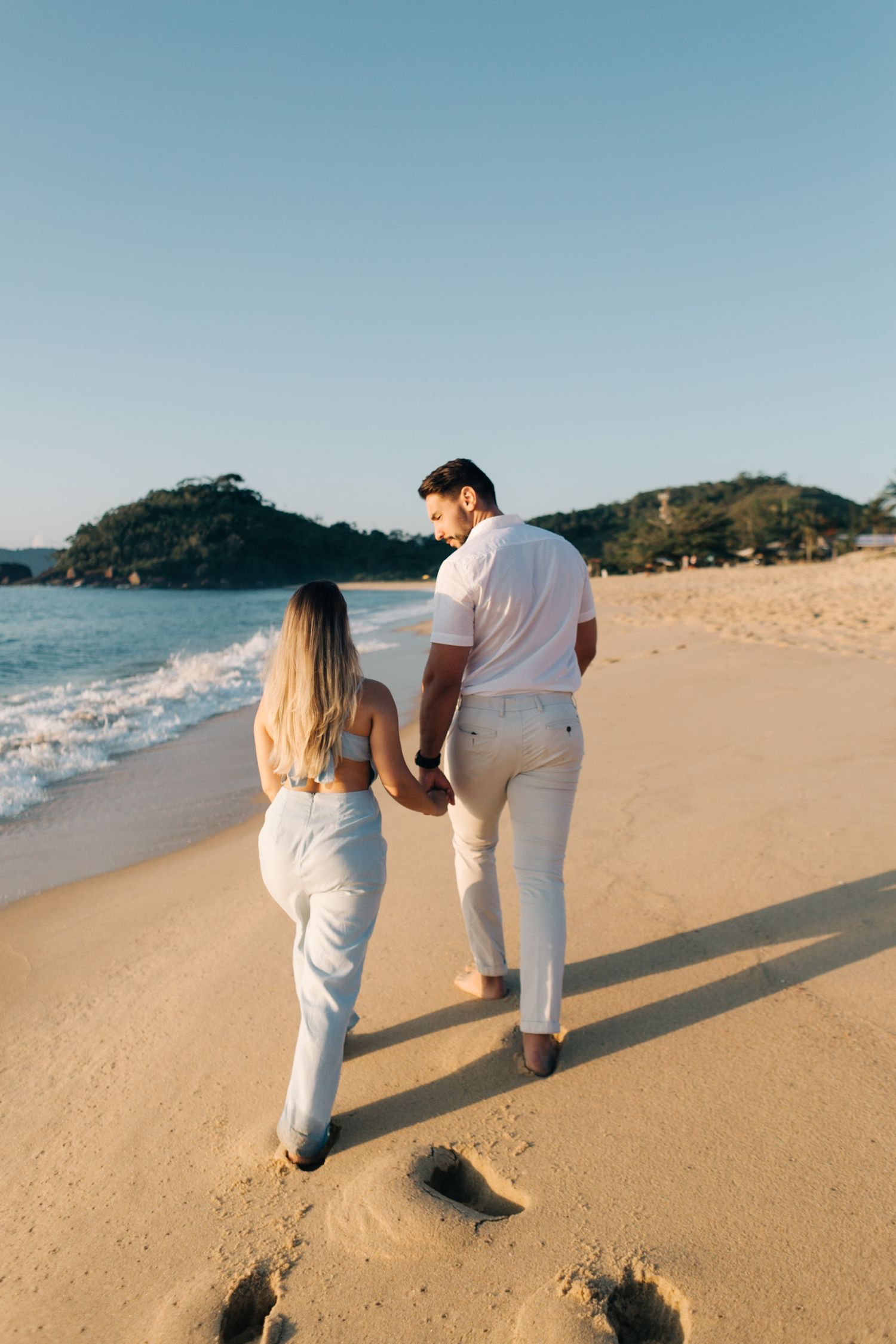Couple in white outfits walking hand in hand on a beach with mountains in the background during sunset.