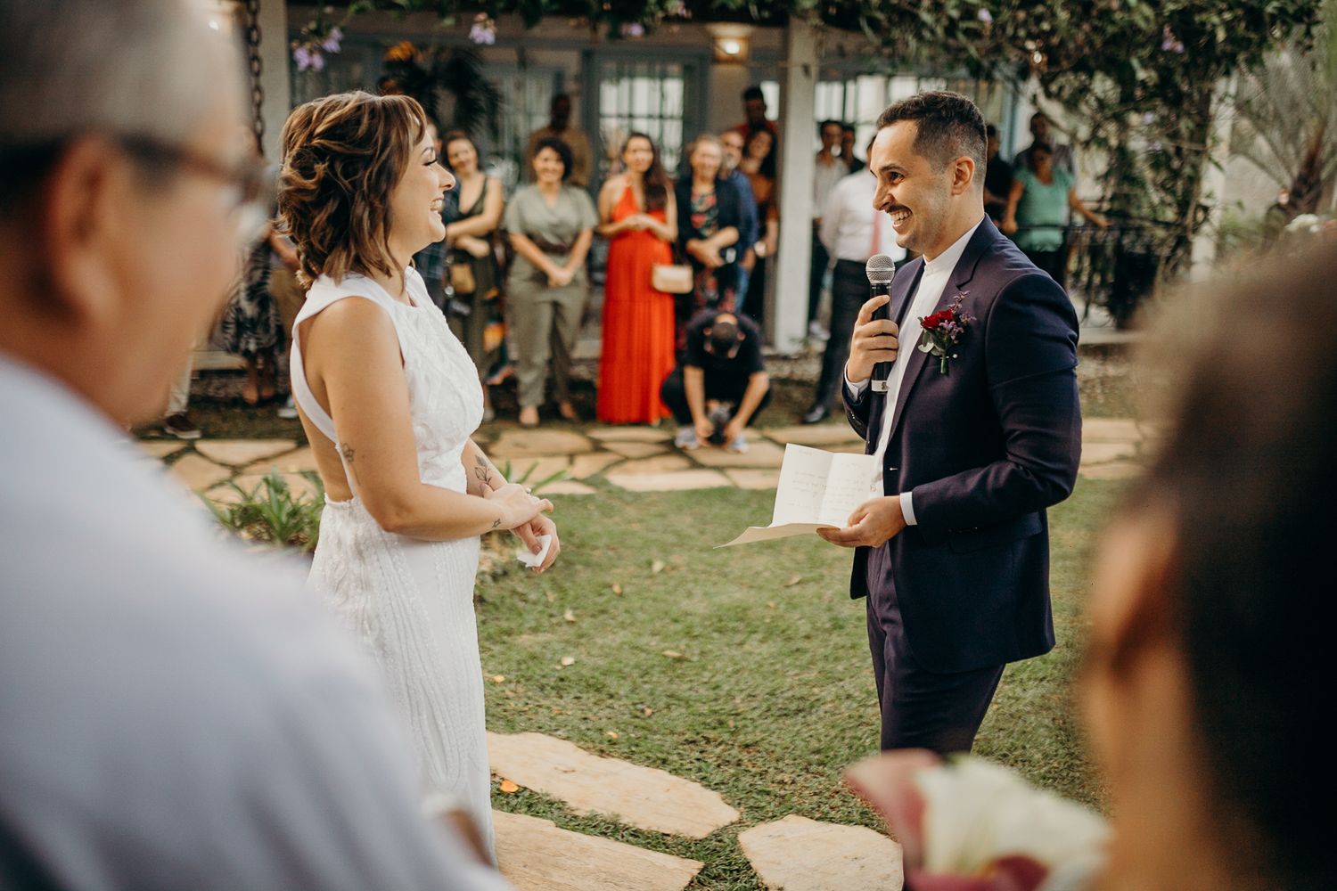 A couple shares their first kiss during an outdoor wedding ceremony as guests look on in a garden setting.