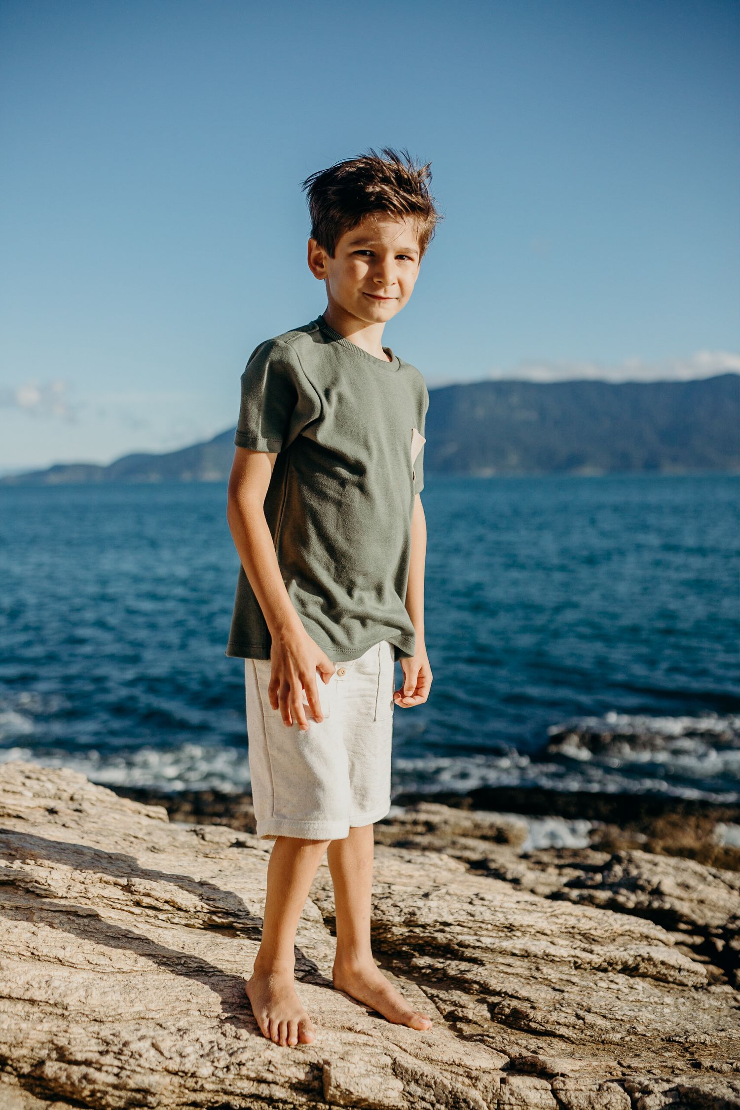 Child stands barefoot on rocky coastal ledge with blue ocean and mountains in background.