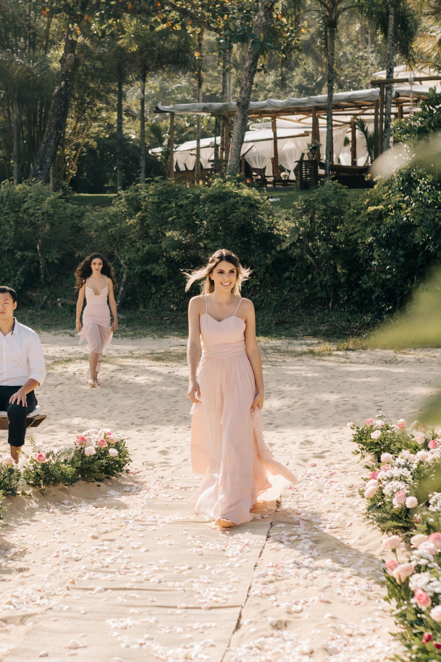 Wedding guests walk along a flower-lined pathway with trees and buildings in the background.