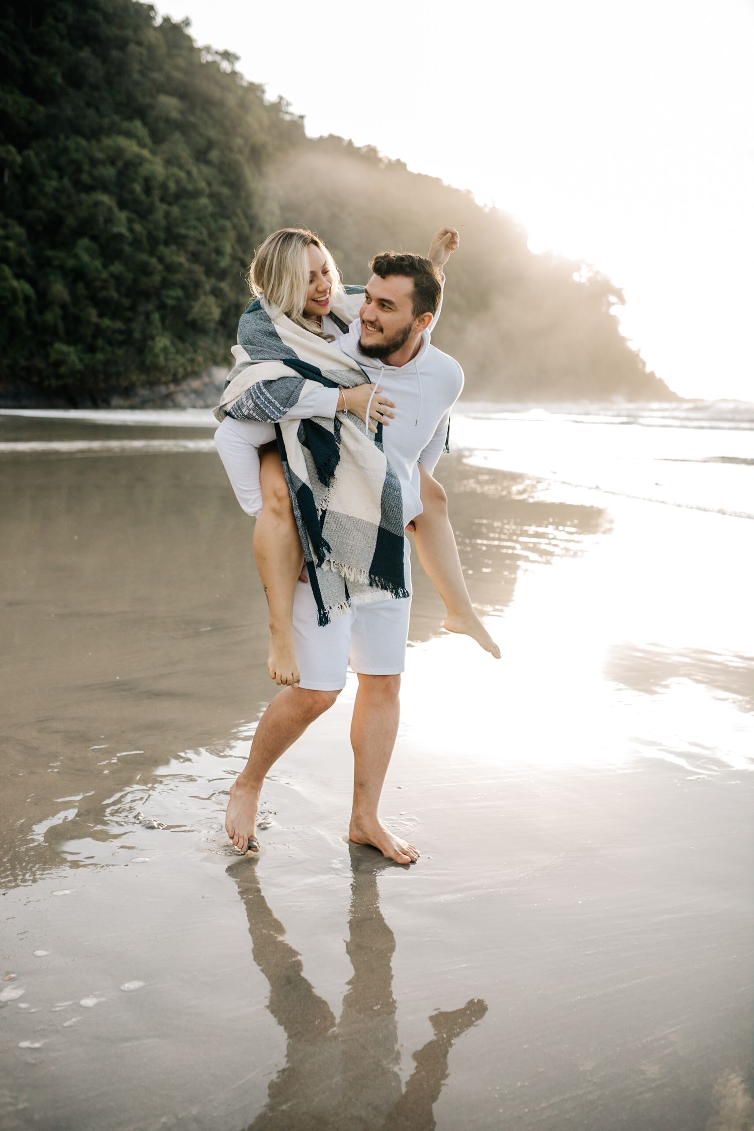 Couple sharing a playful piggyback ride moment on a misty beach at sunset.