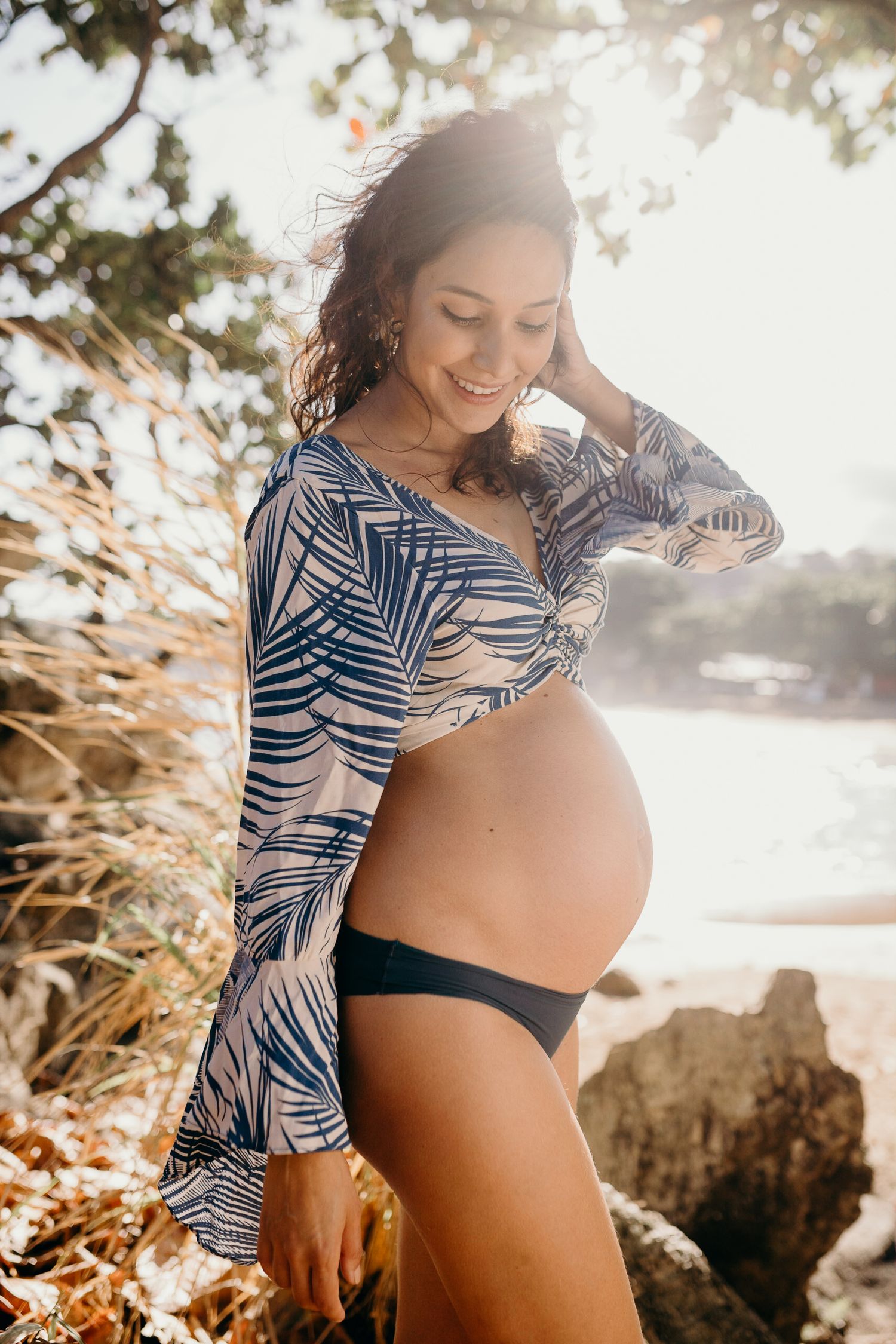 A sun-drenched maternity portrait with tropical foliage and ocean backdrop.