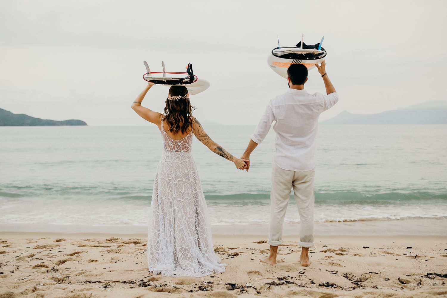 A couple holds surfboards above their heads while walking hand-in-hand on a sandy beach.