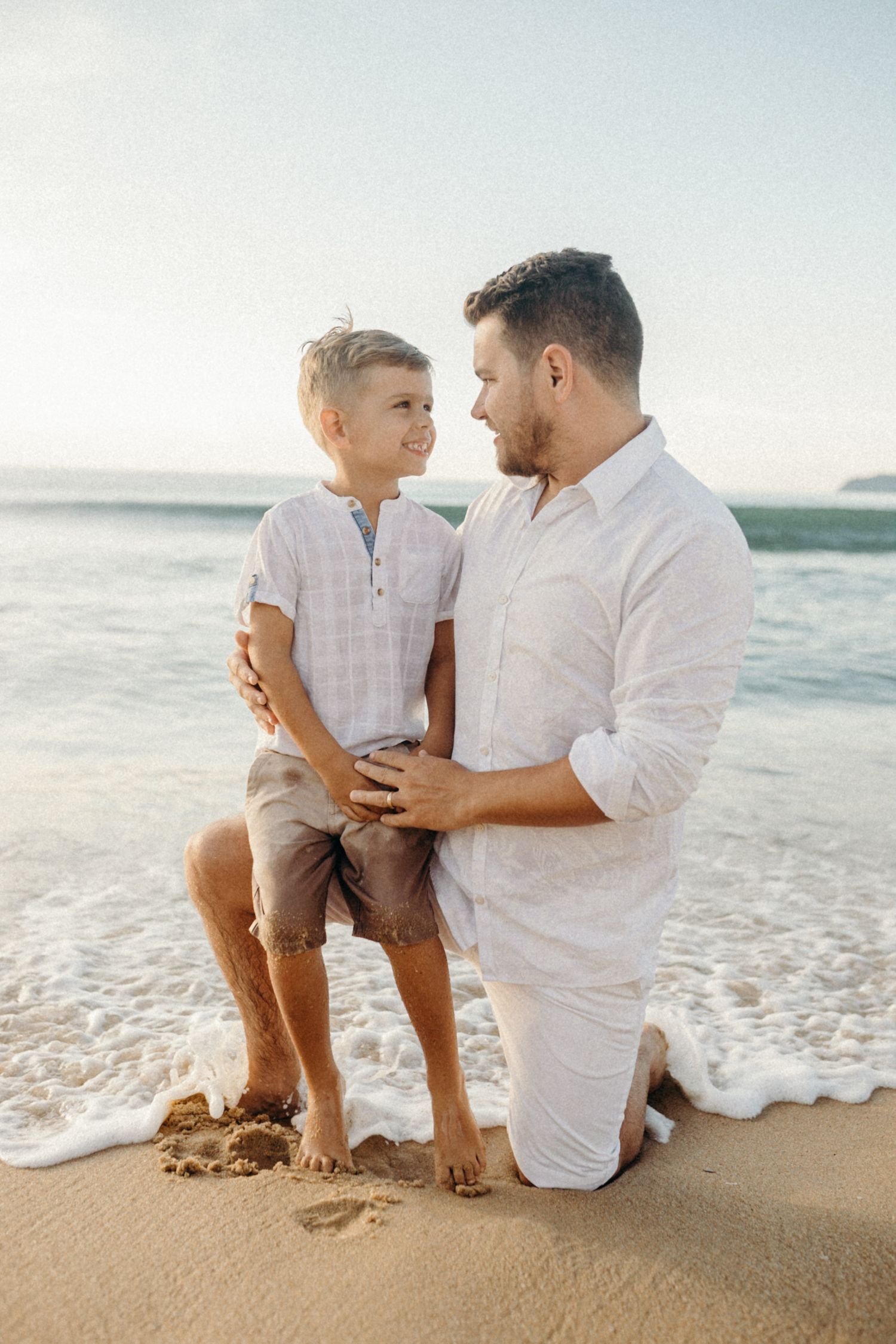 Intimate beachside moment with parent kneeling beside child in matching white shirts.