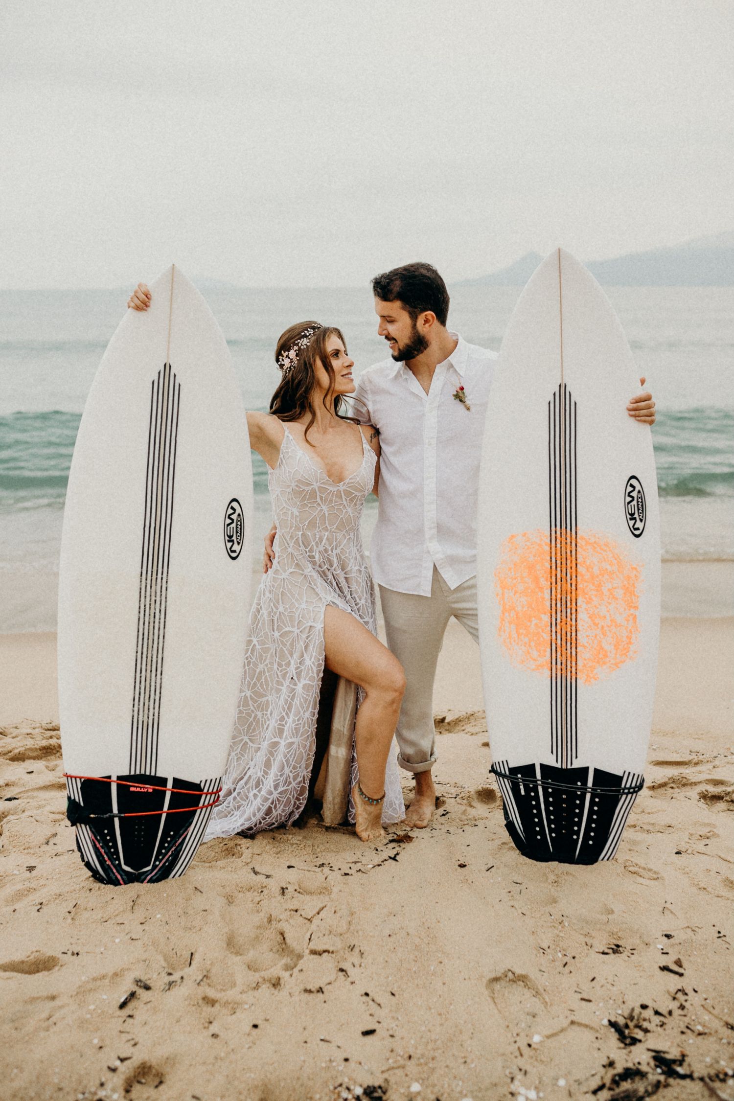Stylish couple poses with surfboards on a beach against a misty ocean backdrop.