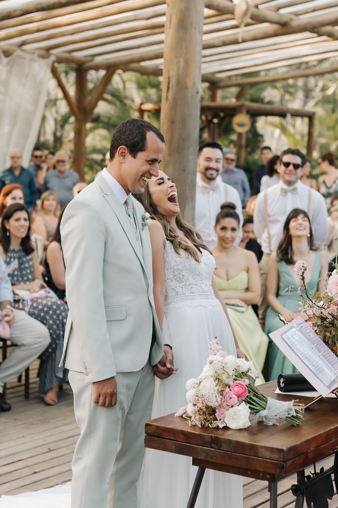 Wedding ceremony with guests gathered under a rustic wooden pergola.