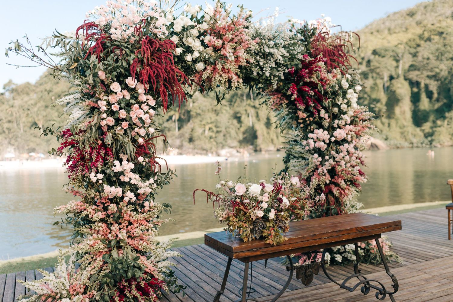 Wedding ceremony arch adorned with pink and burgundy flowers overlooking a lake.