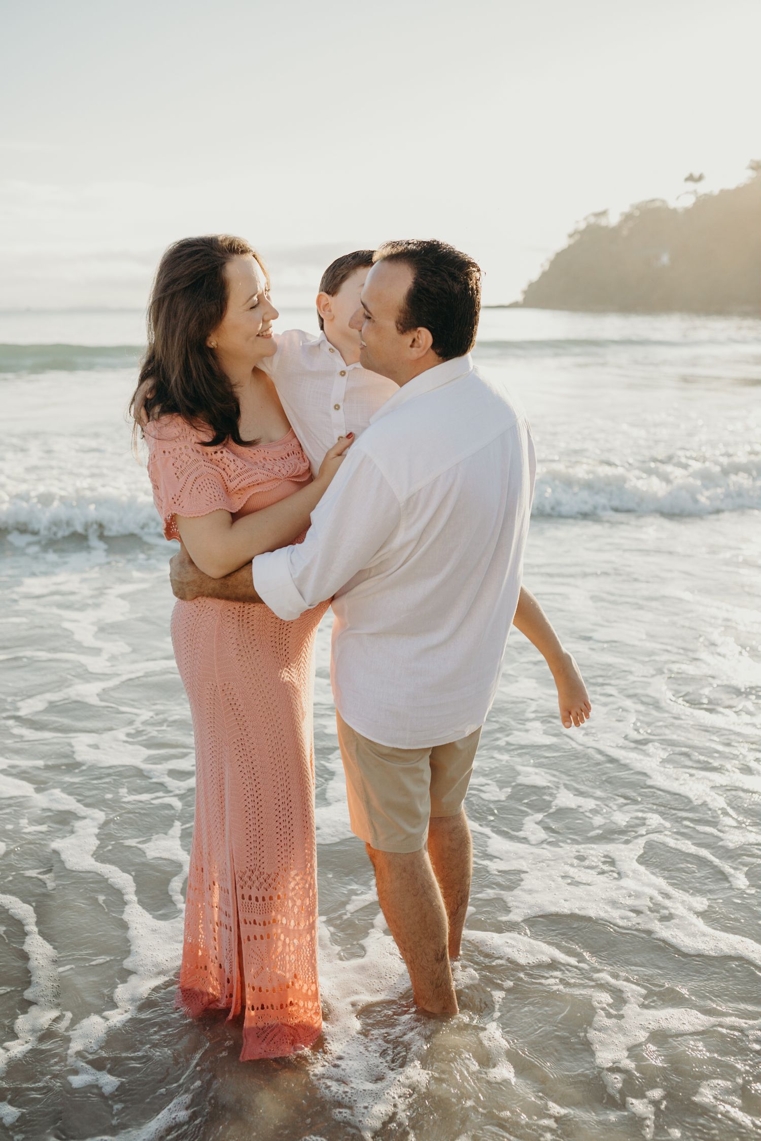 Family shares intimate moments while standing in shallow ocean waves during golden hour.