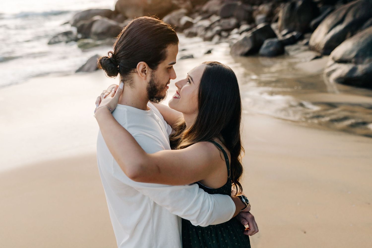 A romantic beach sunset photoshoot of couple embracing on sandy shore with rocky coastline in background.