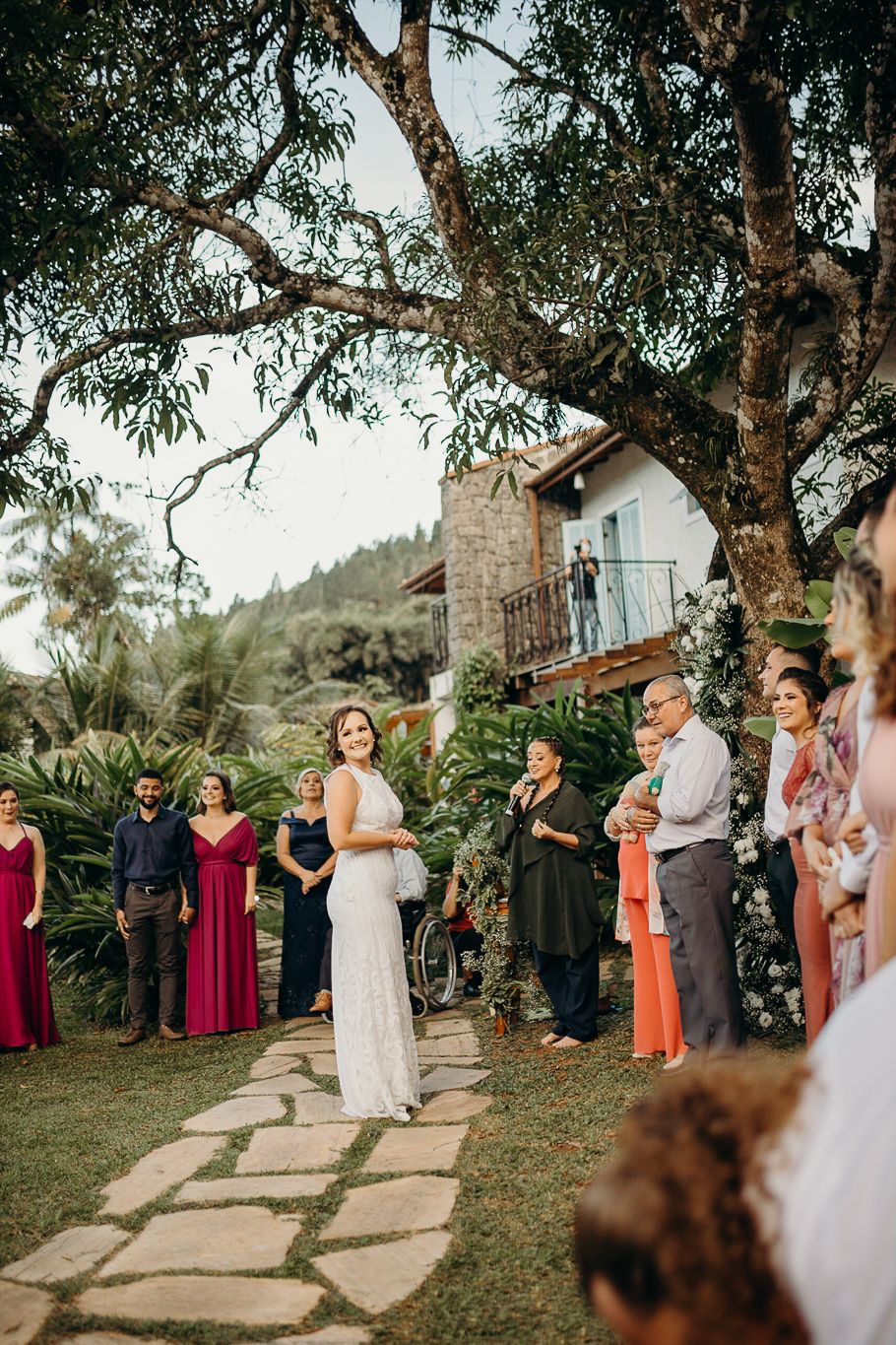 Wedding party gathers on stone pathway with tropical trees and rustic building in background.