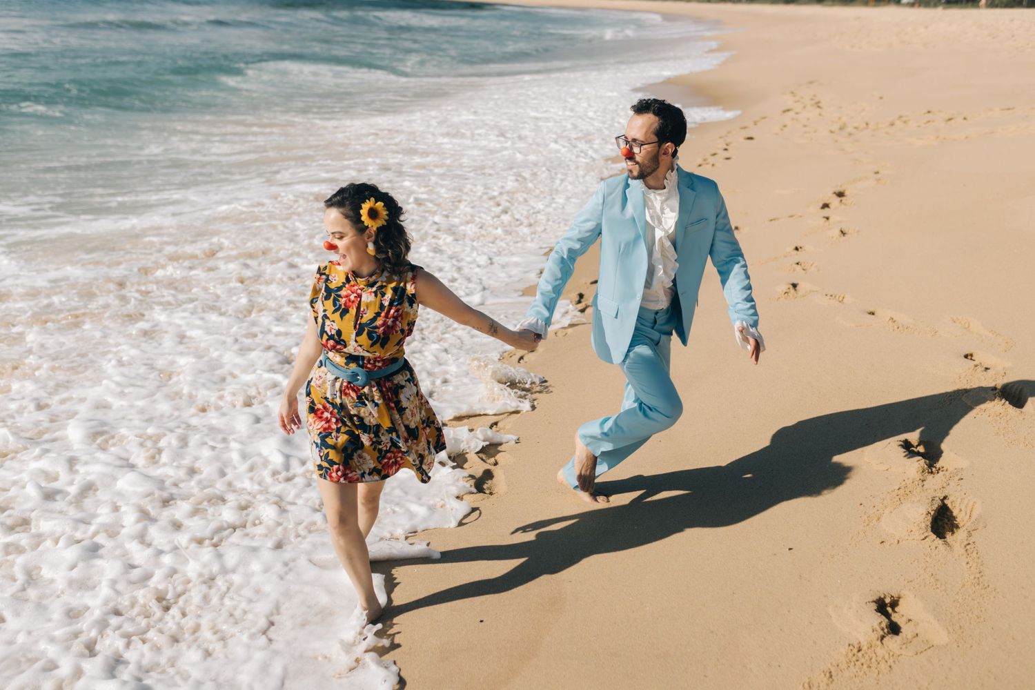 A couple runs playfully along the beach shoreline, wearing a floral dress and light blue suit.