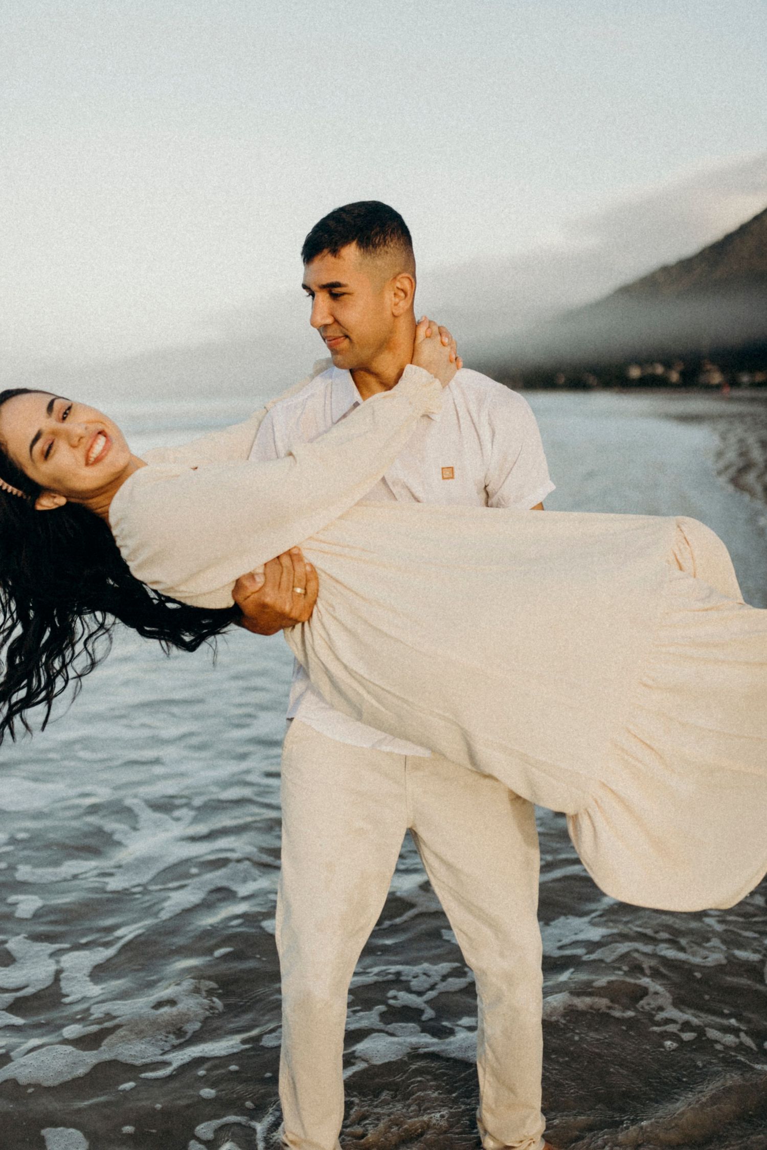 A romantic couple in light-colored outfits share a playful dip pose on a misty beach at sunset.