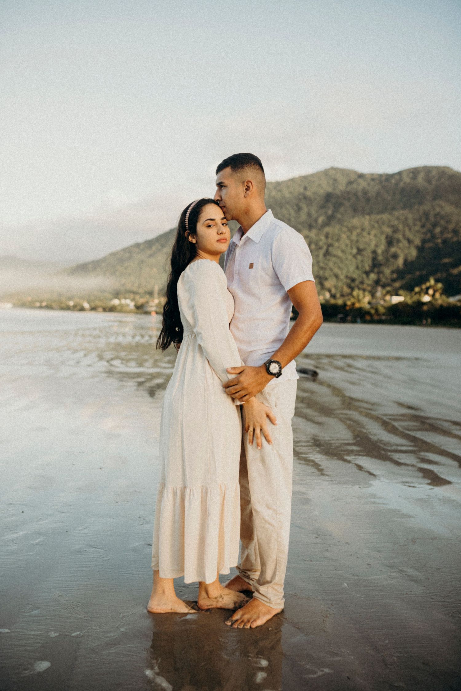 A couple stands together on a beach with mountains and ocean in the background.