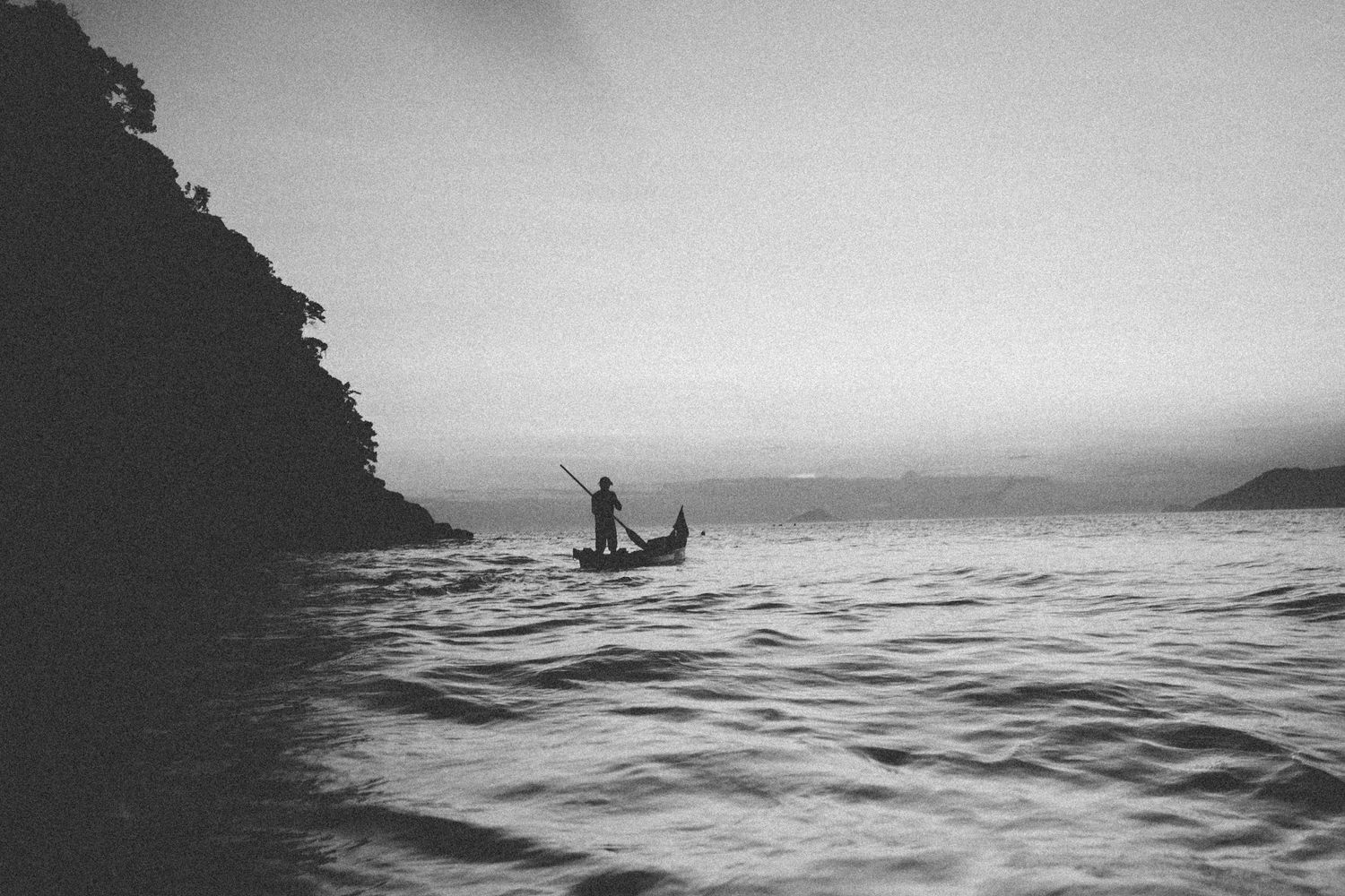 Black and white photo of a lone paddler navigating waves near a rocky coastline.