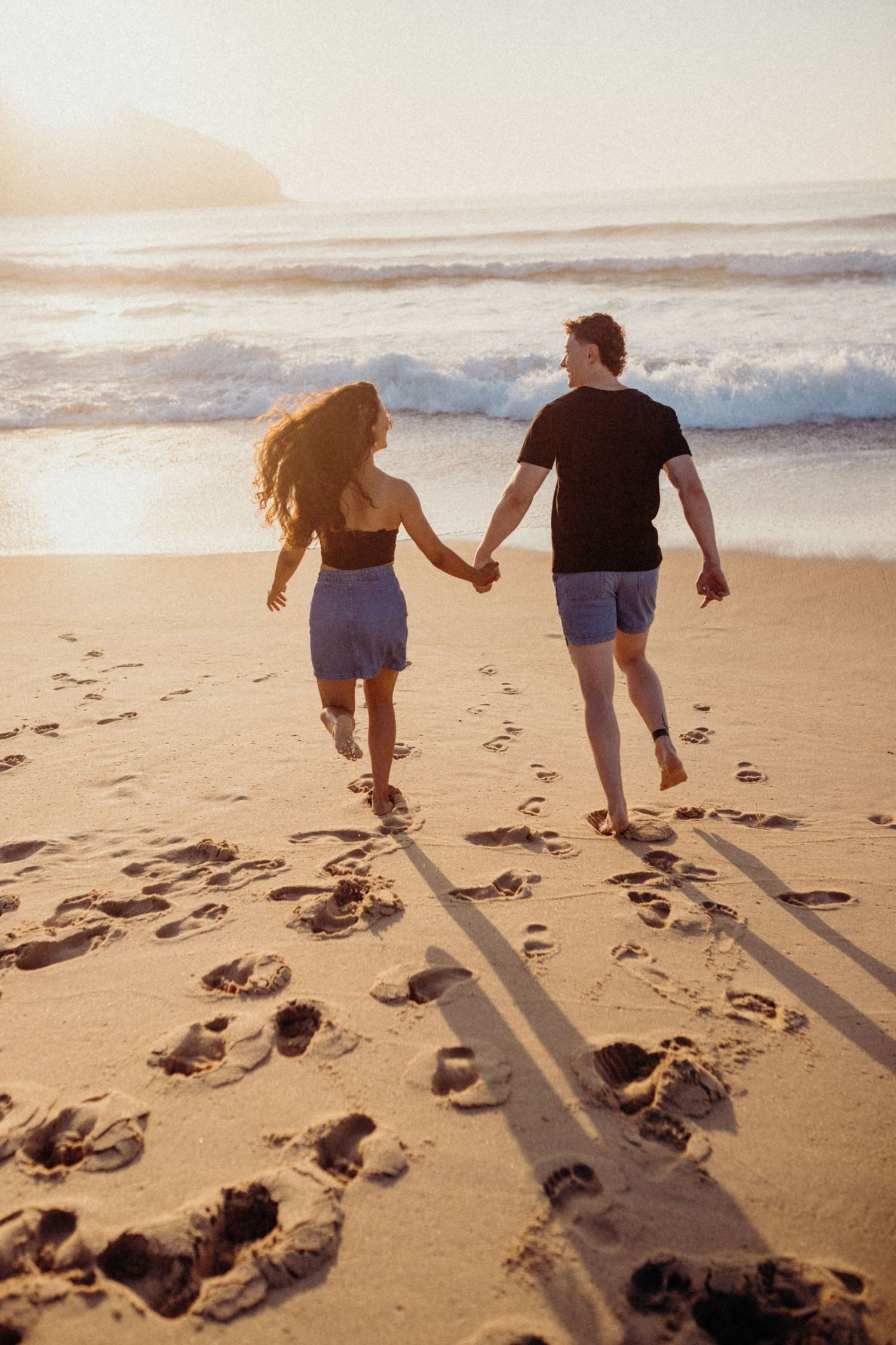 Couple walks hand in hand on beach leaving footprints in sand during golden hour.
