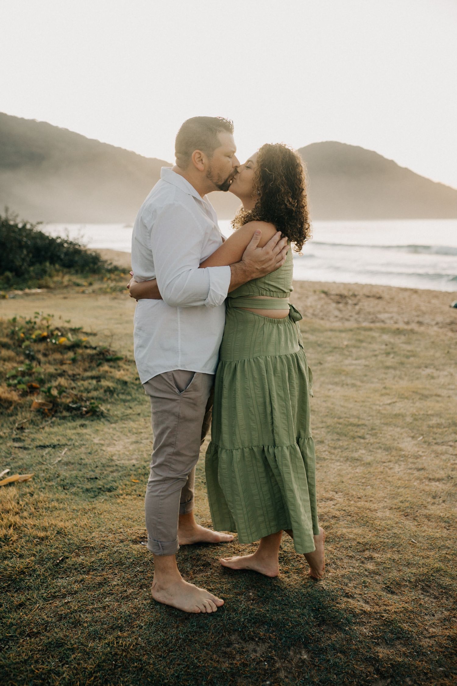 Romantic couple embracing on grassy cliff overlooking ocean at sunset.