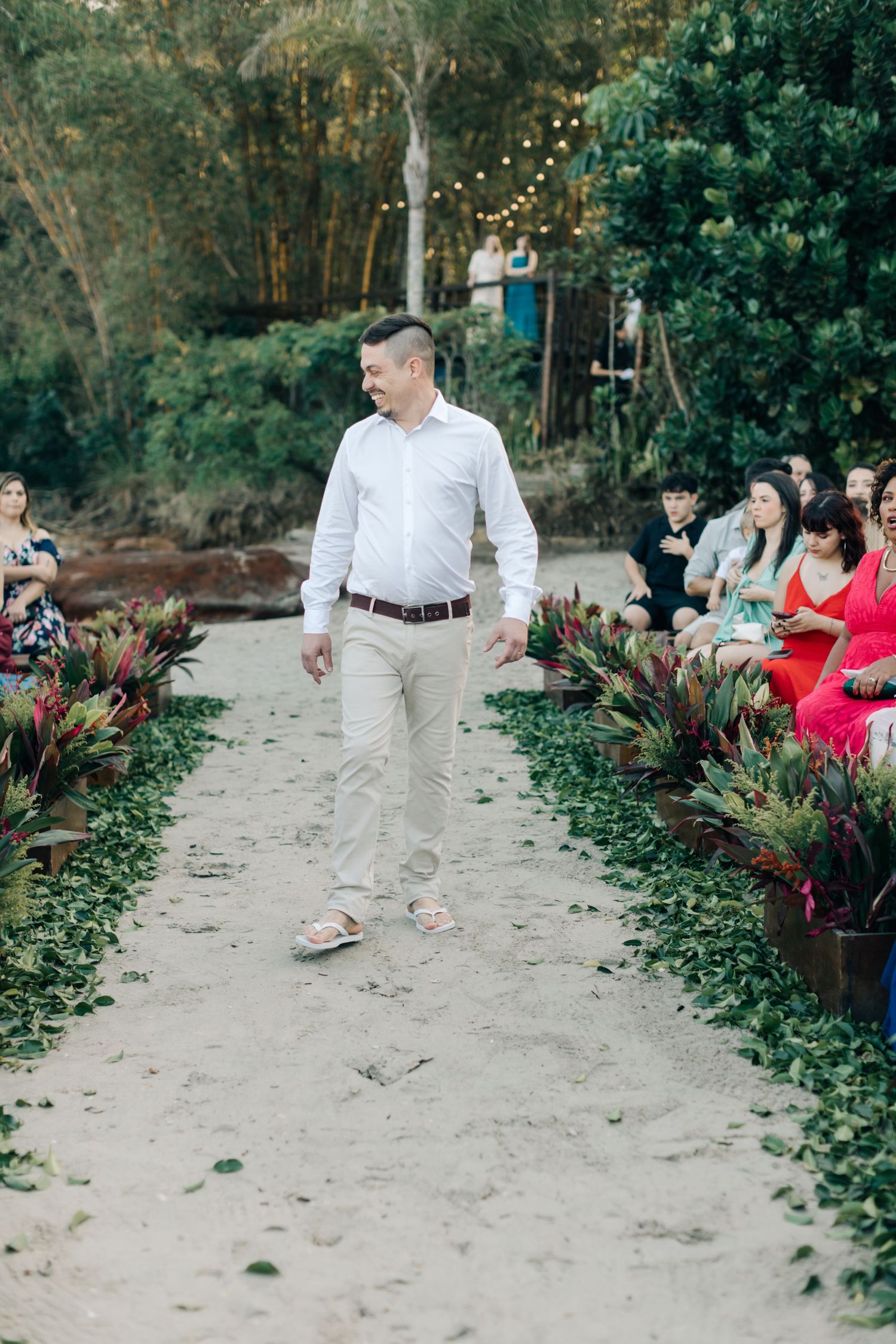 Guest walking down sandy aisle lined with potted plants at outdoor ceremony.