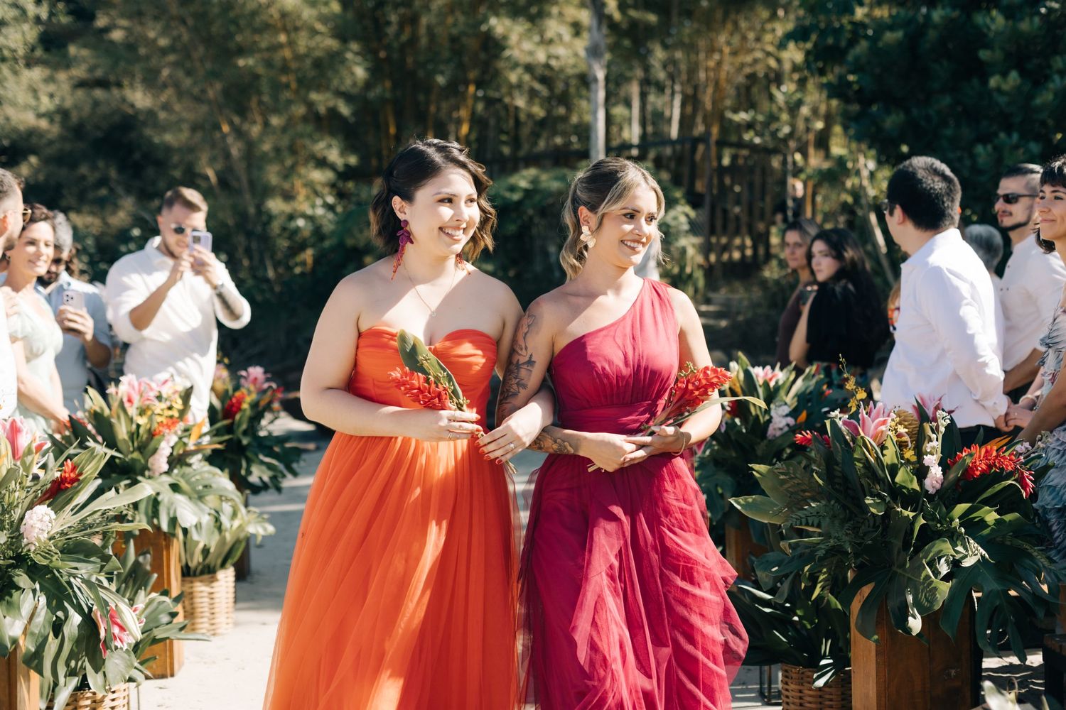 Bridesmaids in vibrant orange and red dresses walk along a tropical garden path during an outdoor wedding ceremony.