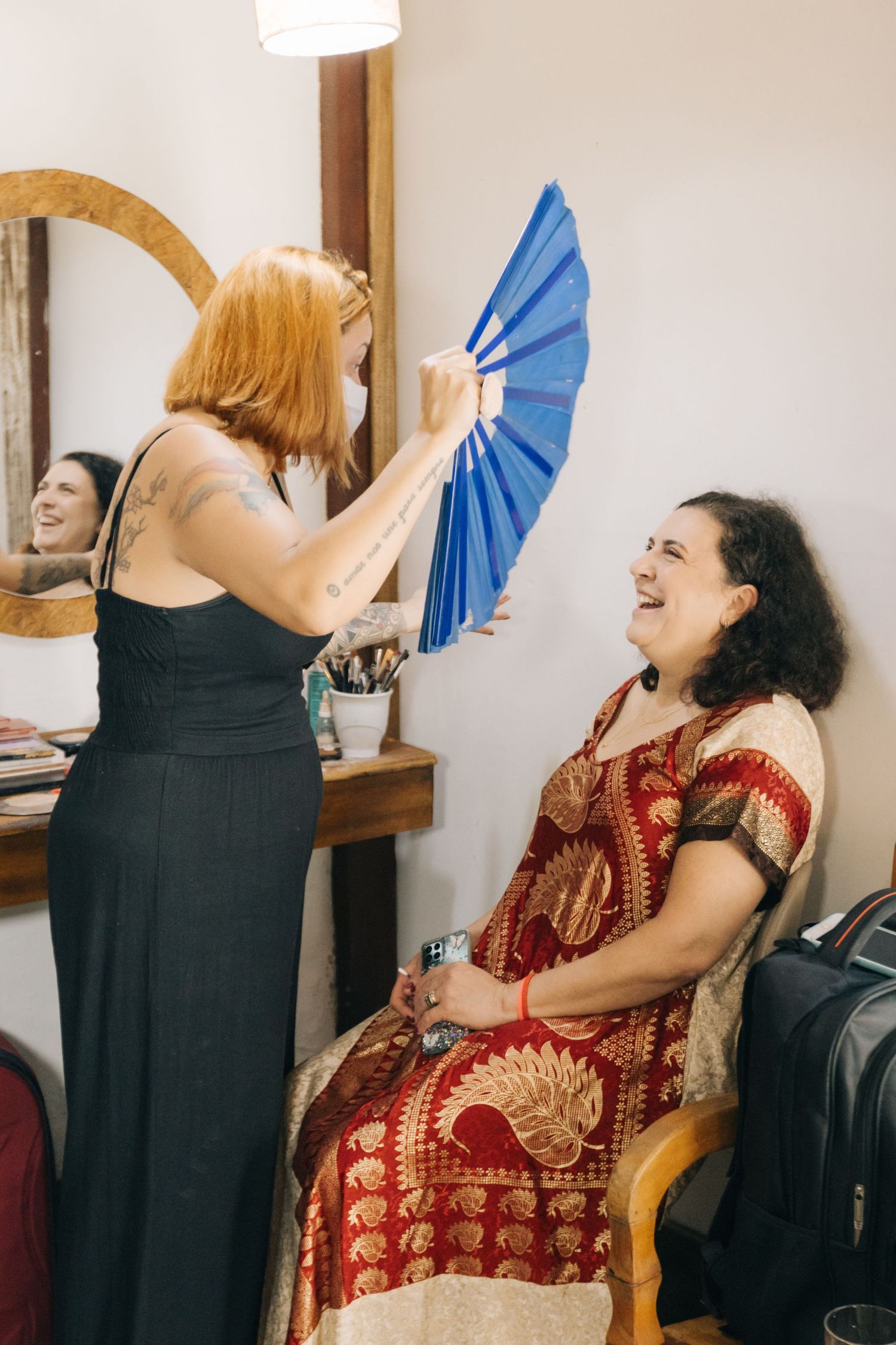 A person in a red dress smiles and holds a bright blue fan while getting ready.