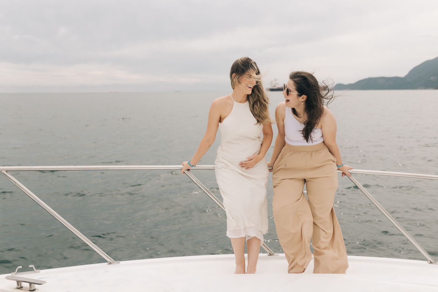 Two people in light colored outfits chat while standing on a boat deck.