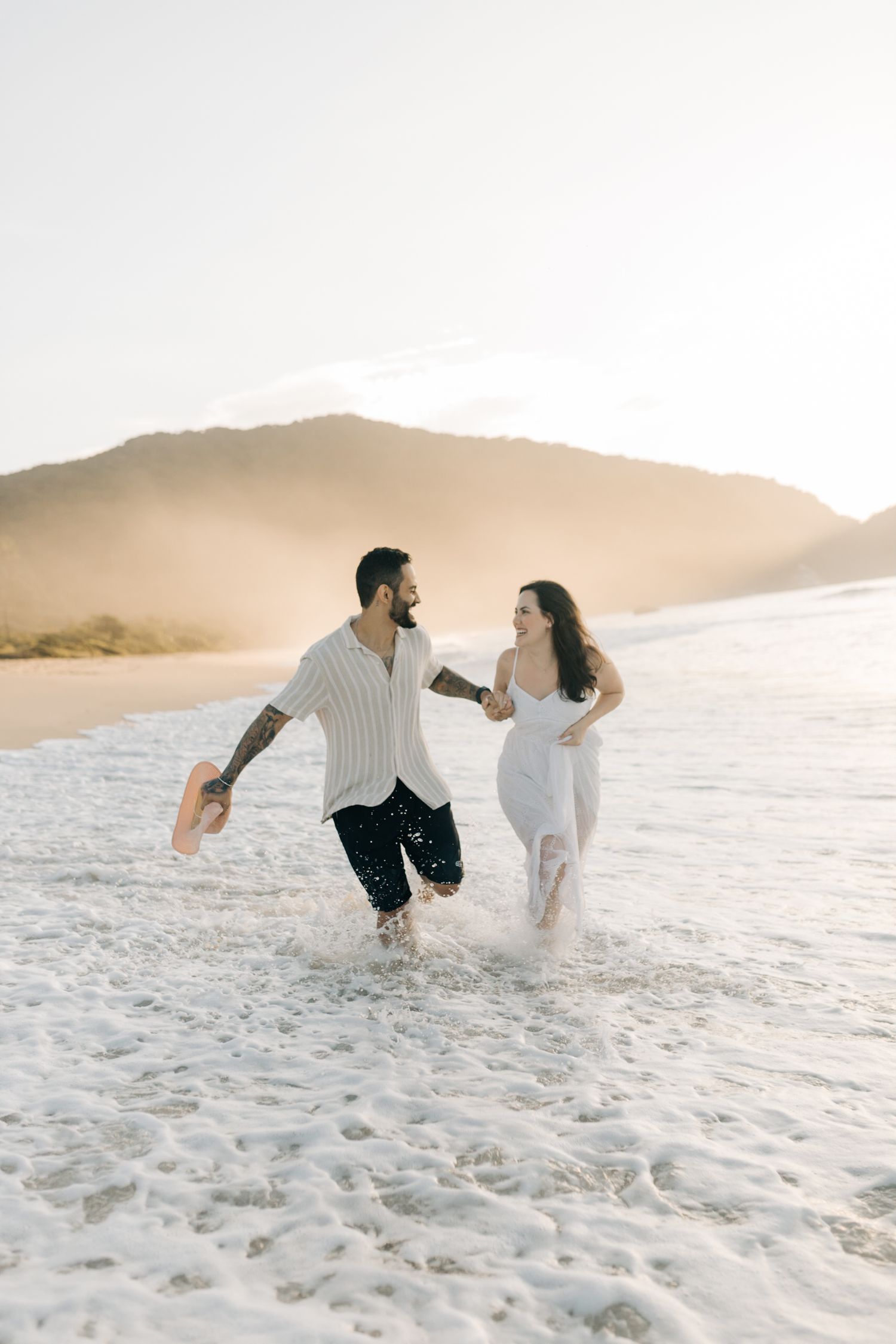 Two people playfully run through ocean waves at a beach during golden hour.