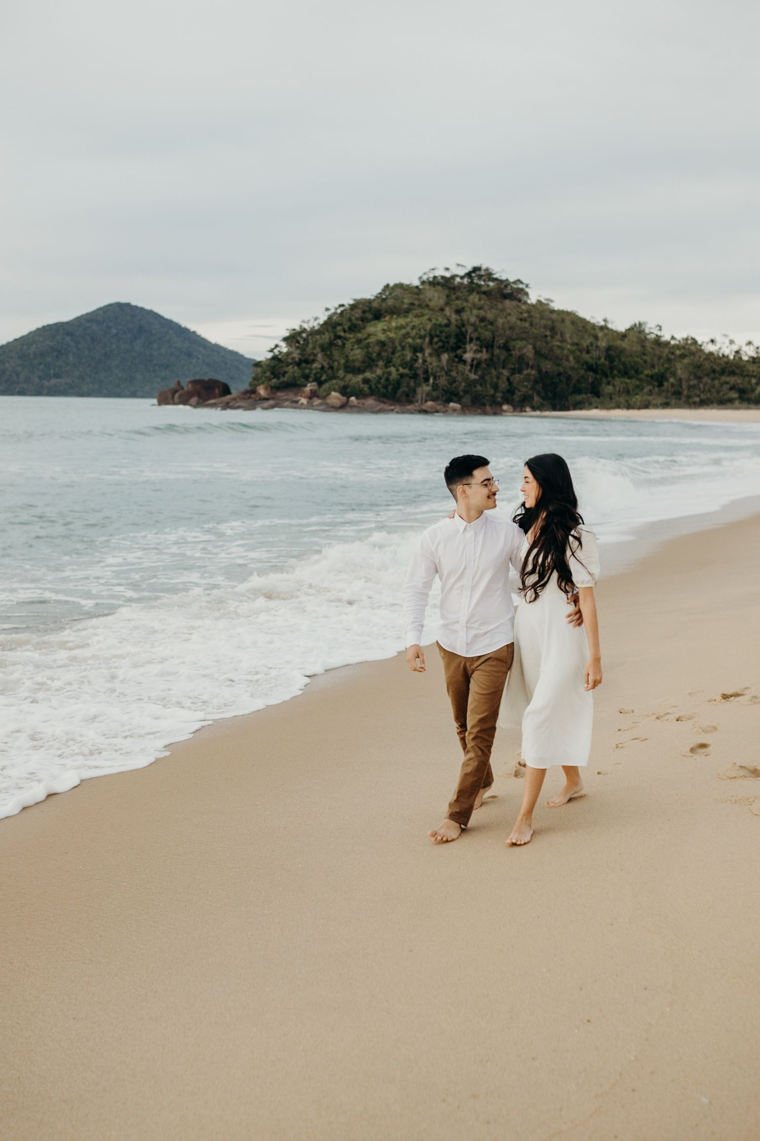 A couple strolls hand in hand along a scenic beach with mountains in the background at sunset.