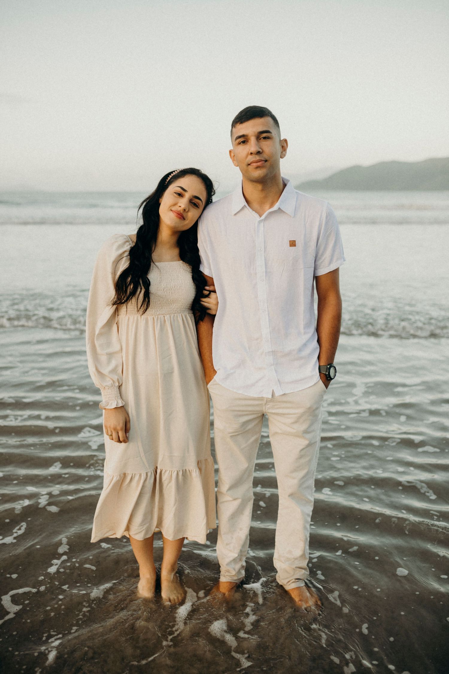 Two people in coordinating cream and white outfits pose together on a dark sandy beach.
