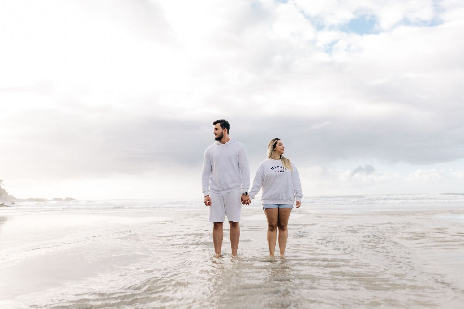 Couple in coordinated white outfits walking together in shallow ocean water.