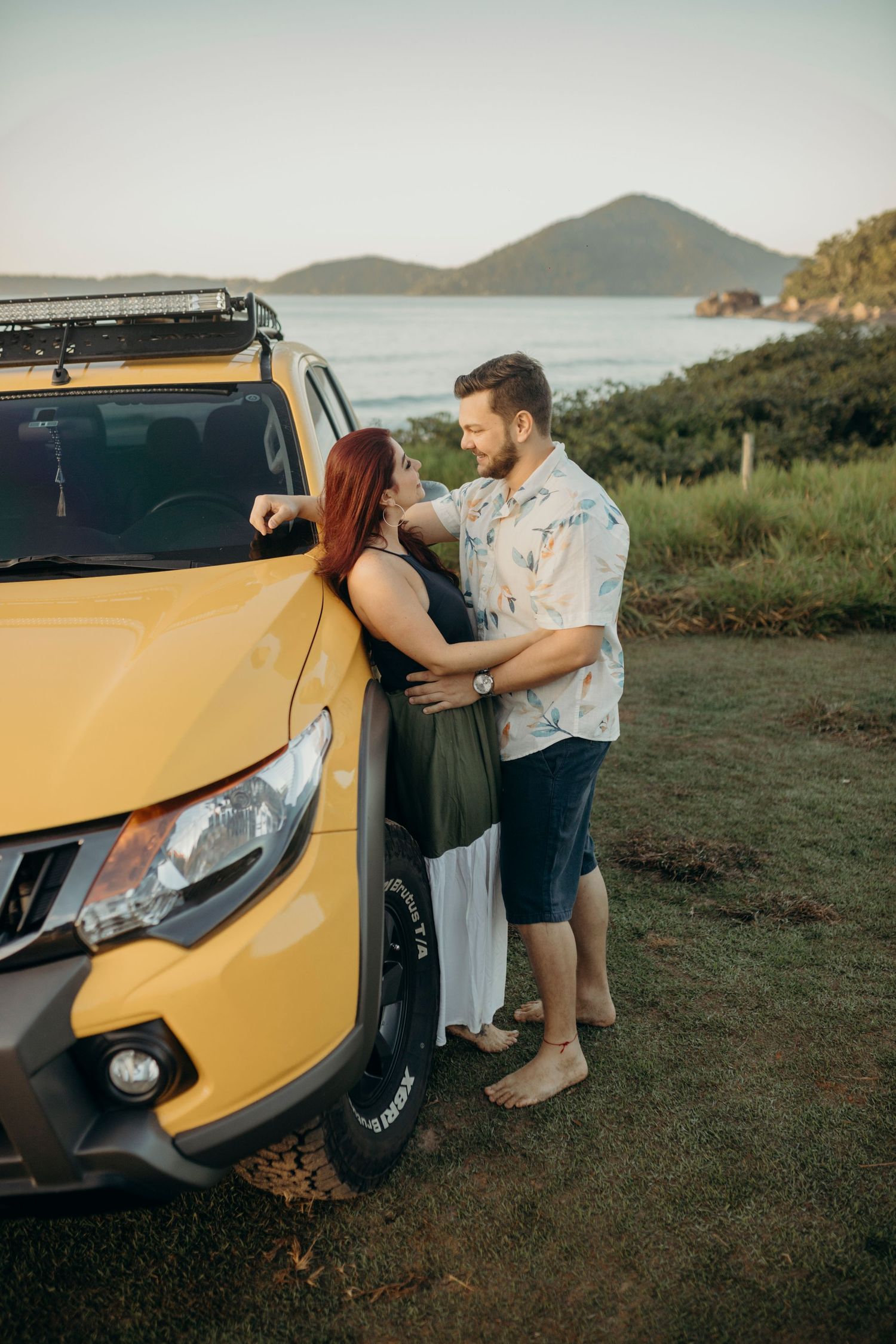 Couple standing beside yellow pickup truck with ocean view and mountains in background.