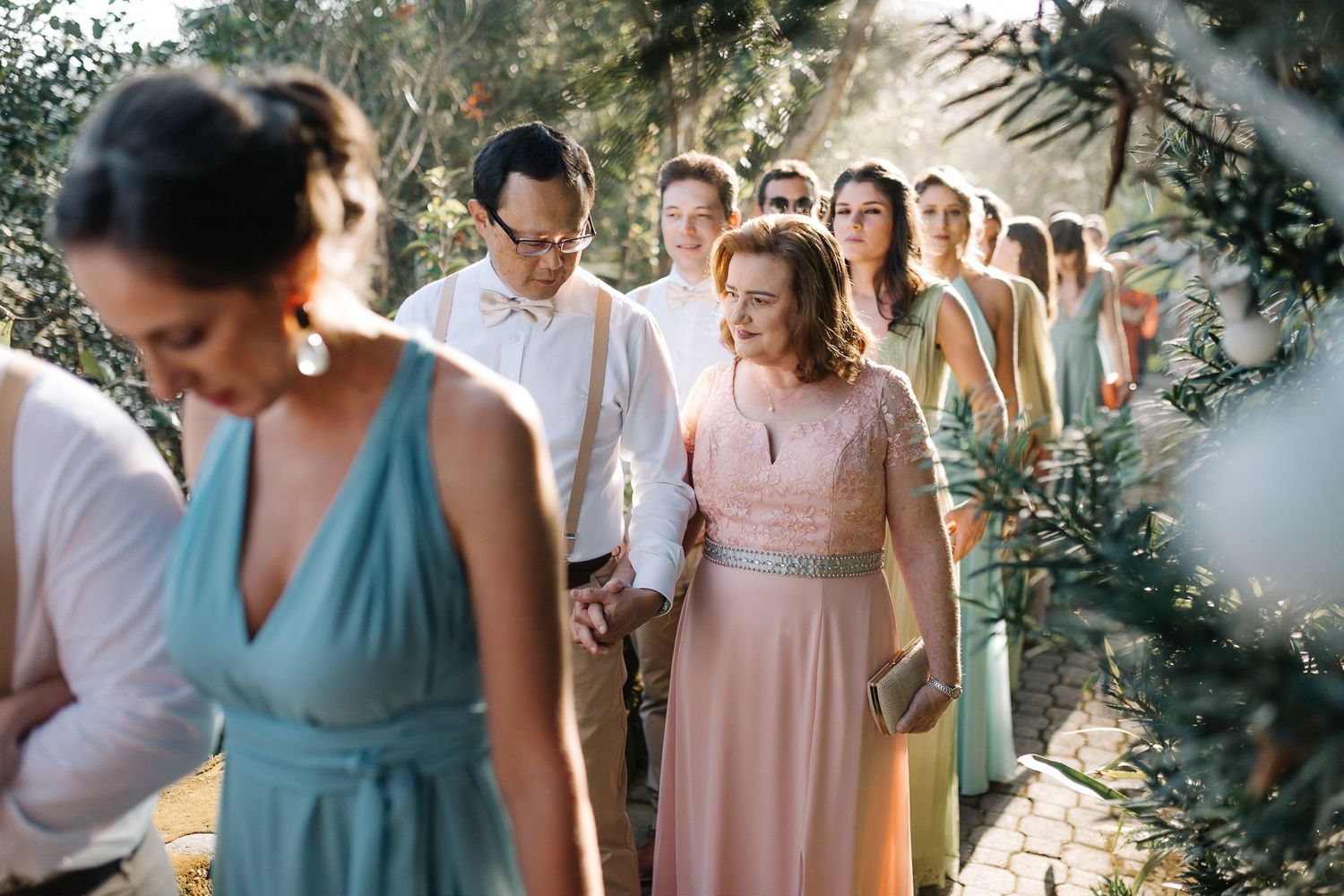 Wedding party members in pastel dresses walking in line along outdoor garden path.