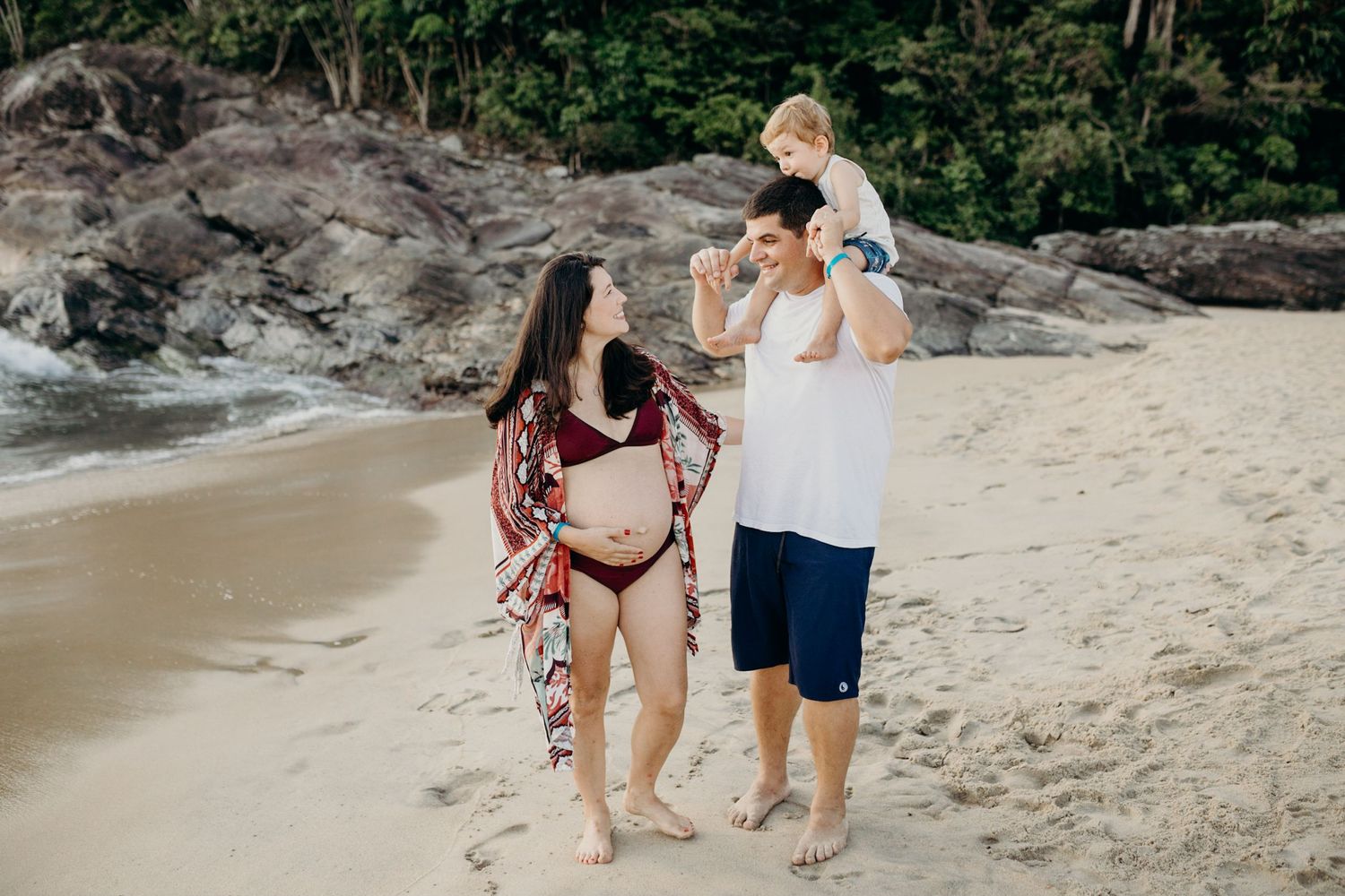 Family walking along a scenic beach with rocky outcrops and lush forest scenery.