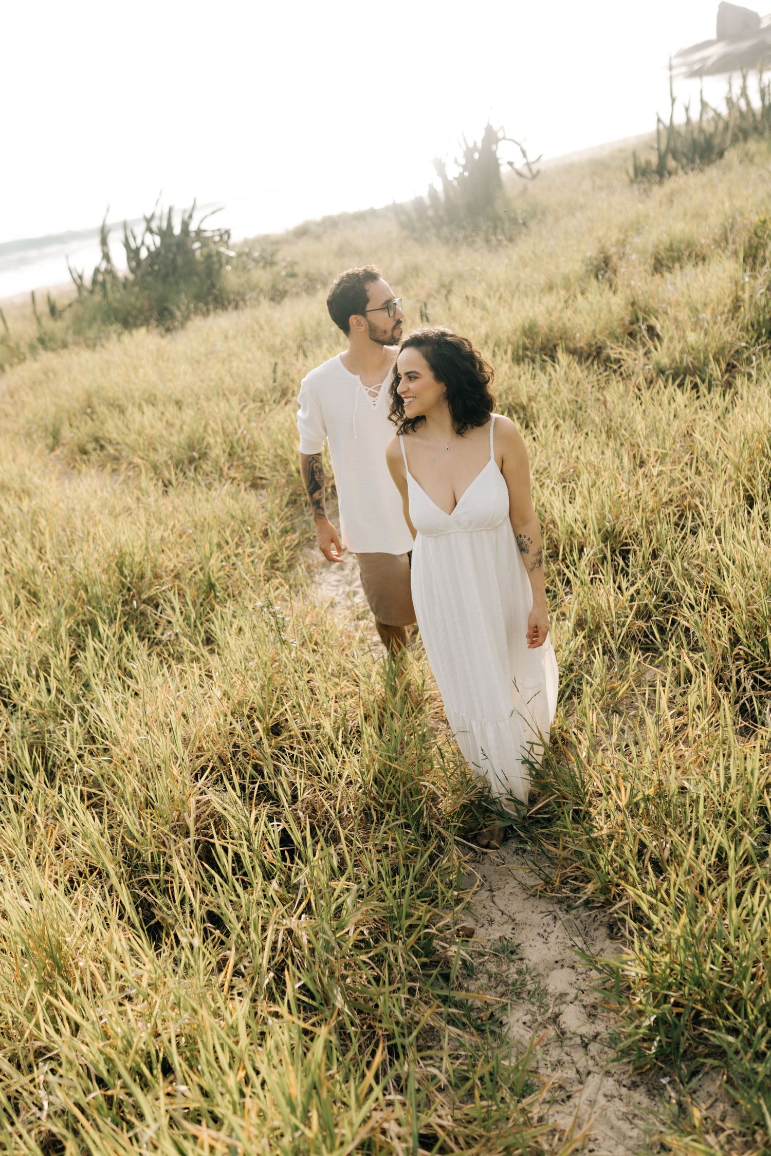 Couple in white outfits walking through tall grass field at sunset.