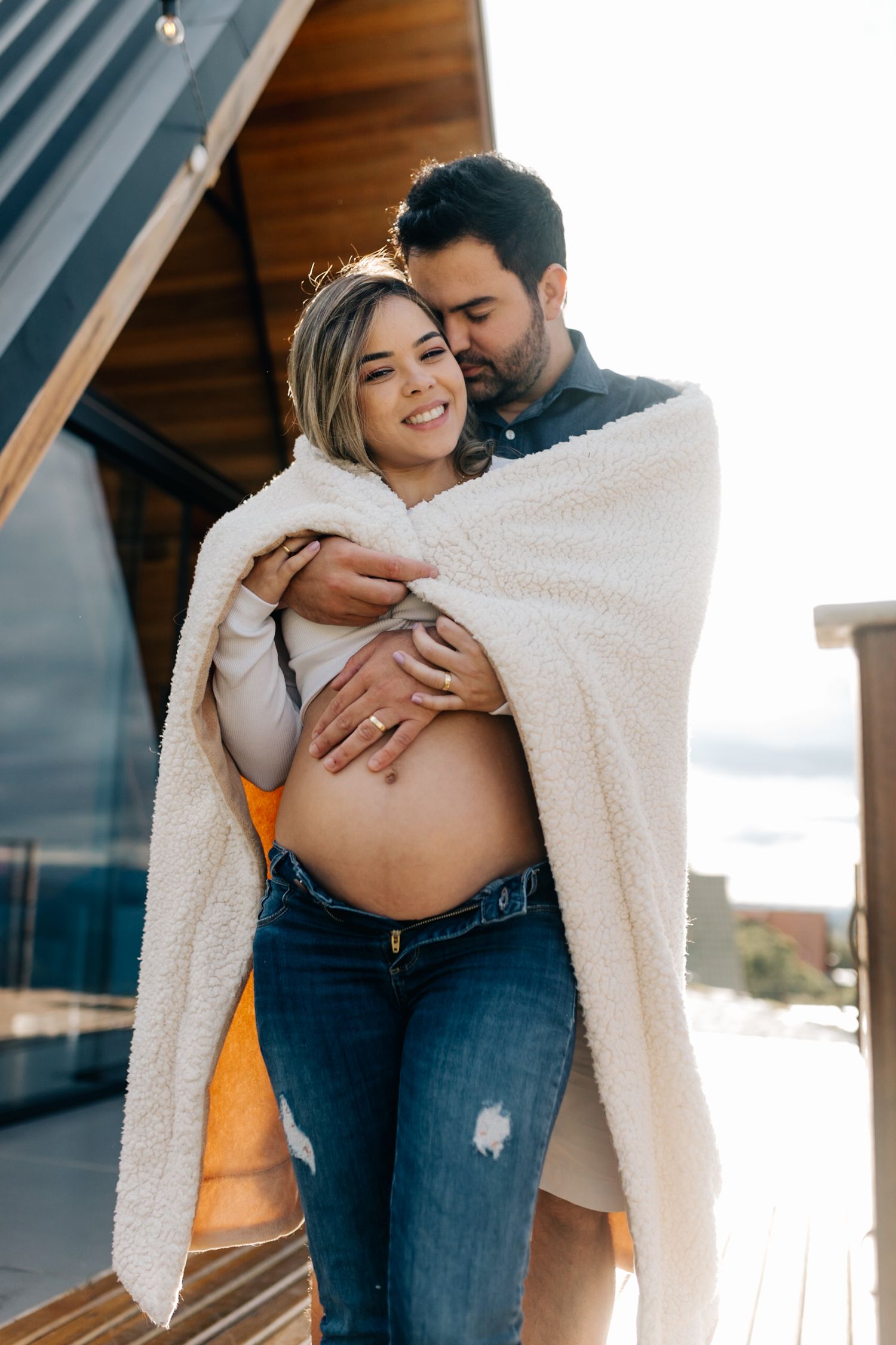 Intimate maternity moment wrapped in a soft white blanket outside an A-frame cabin.