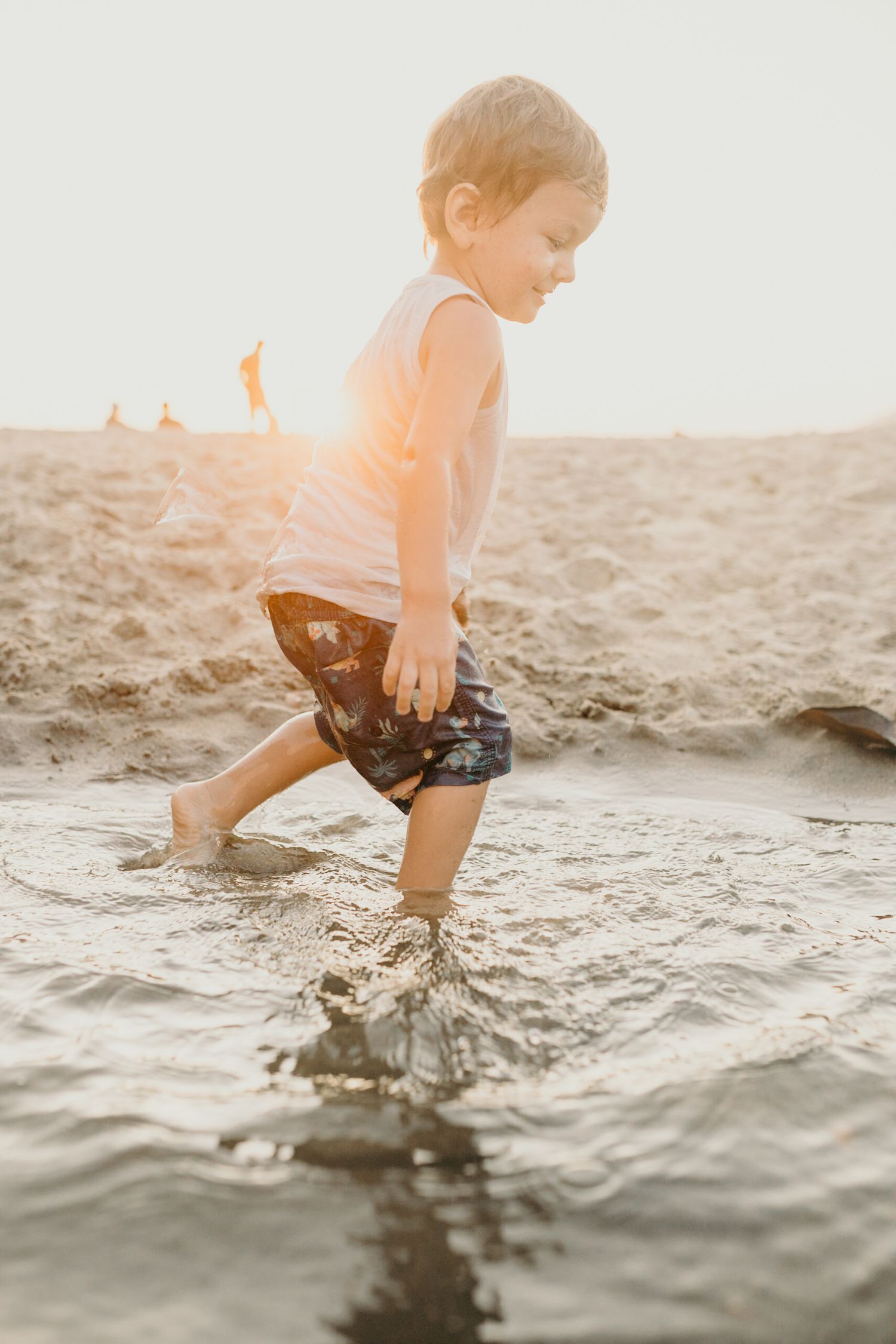 Young child in wet clothes walks through shallow beach water during golden hour.