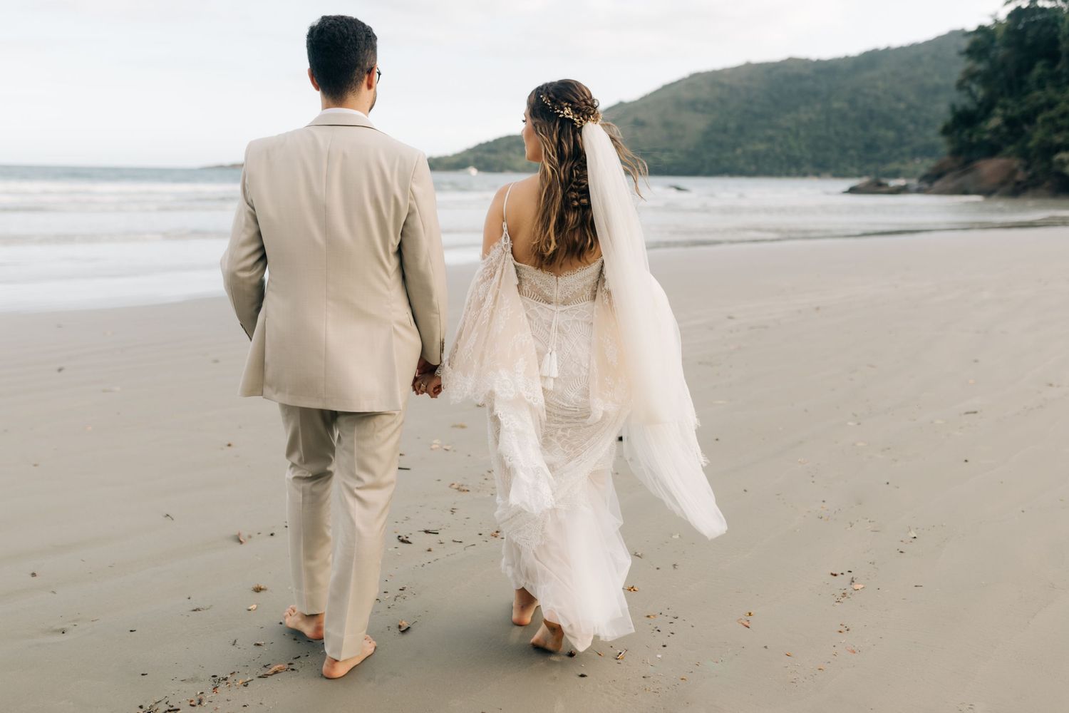 Newlyweds walking barefoot on beach with flowing wedding dress and veil trailing behind.