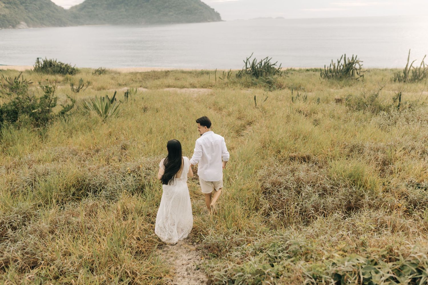A couple walks along a coastal path wearing white outfits with ocean views in the background.