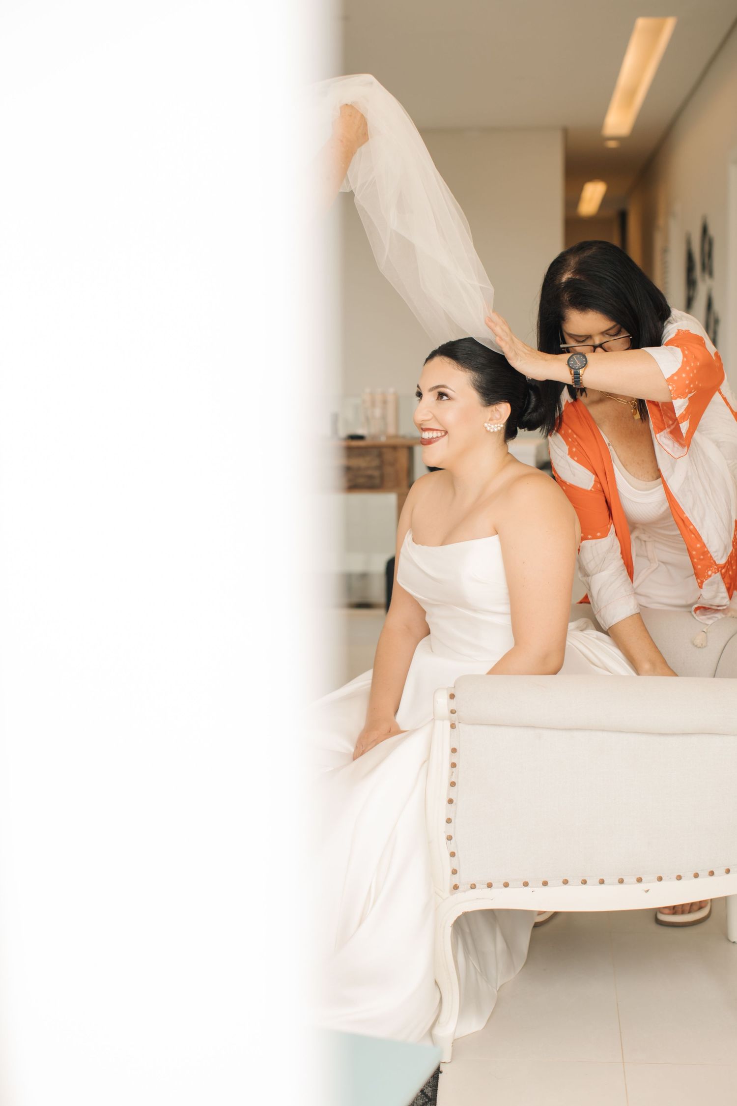 Wedding preparation moment captured with person in white gown seated on upholstered bench.