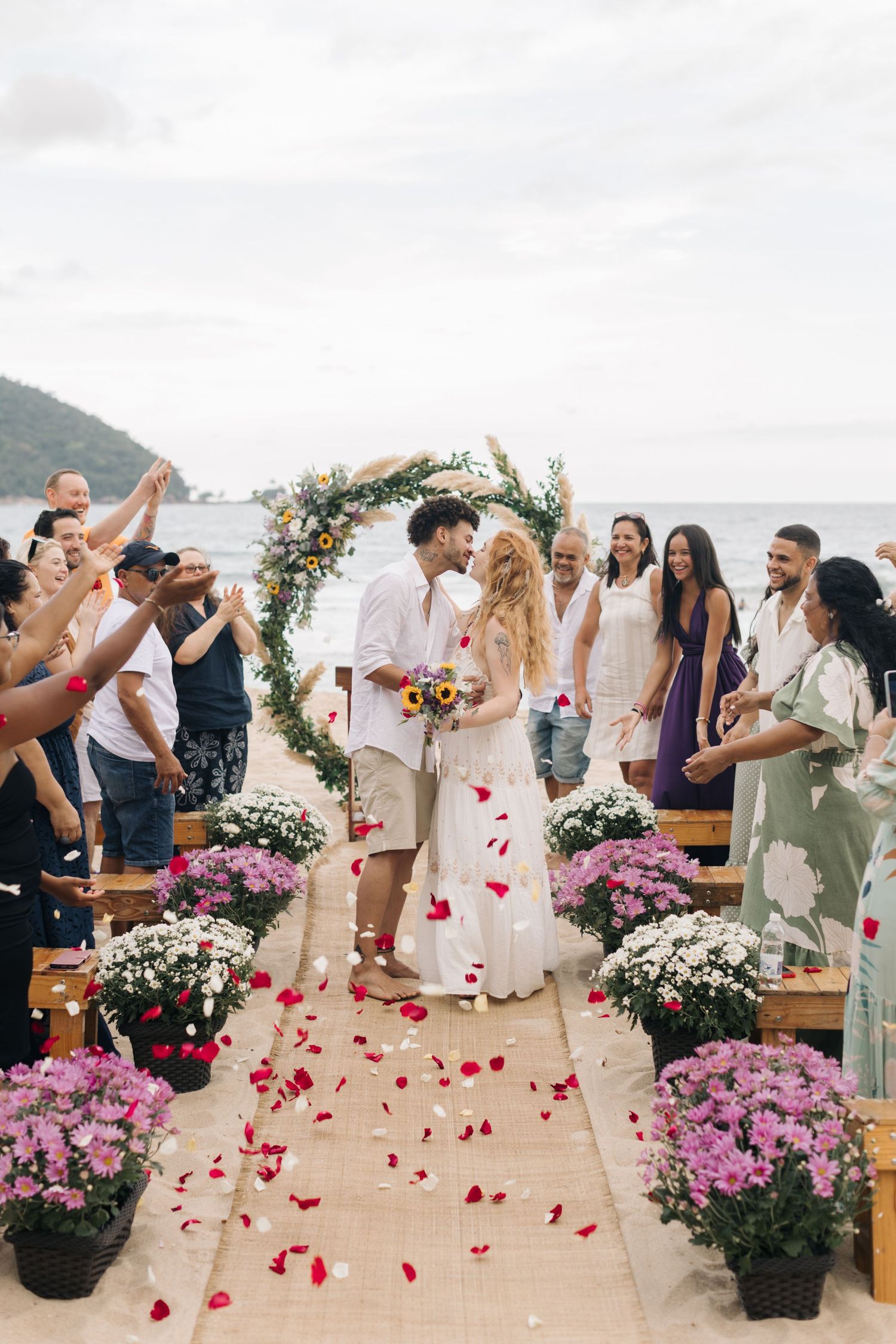 Guests celebrate and throw rose petals at a beach wedding with pink floral decorations lining the aisle.
