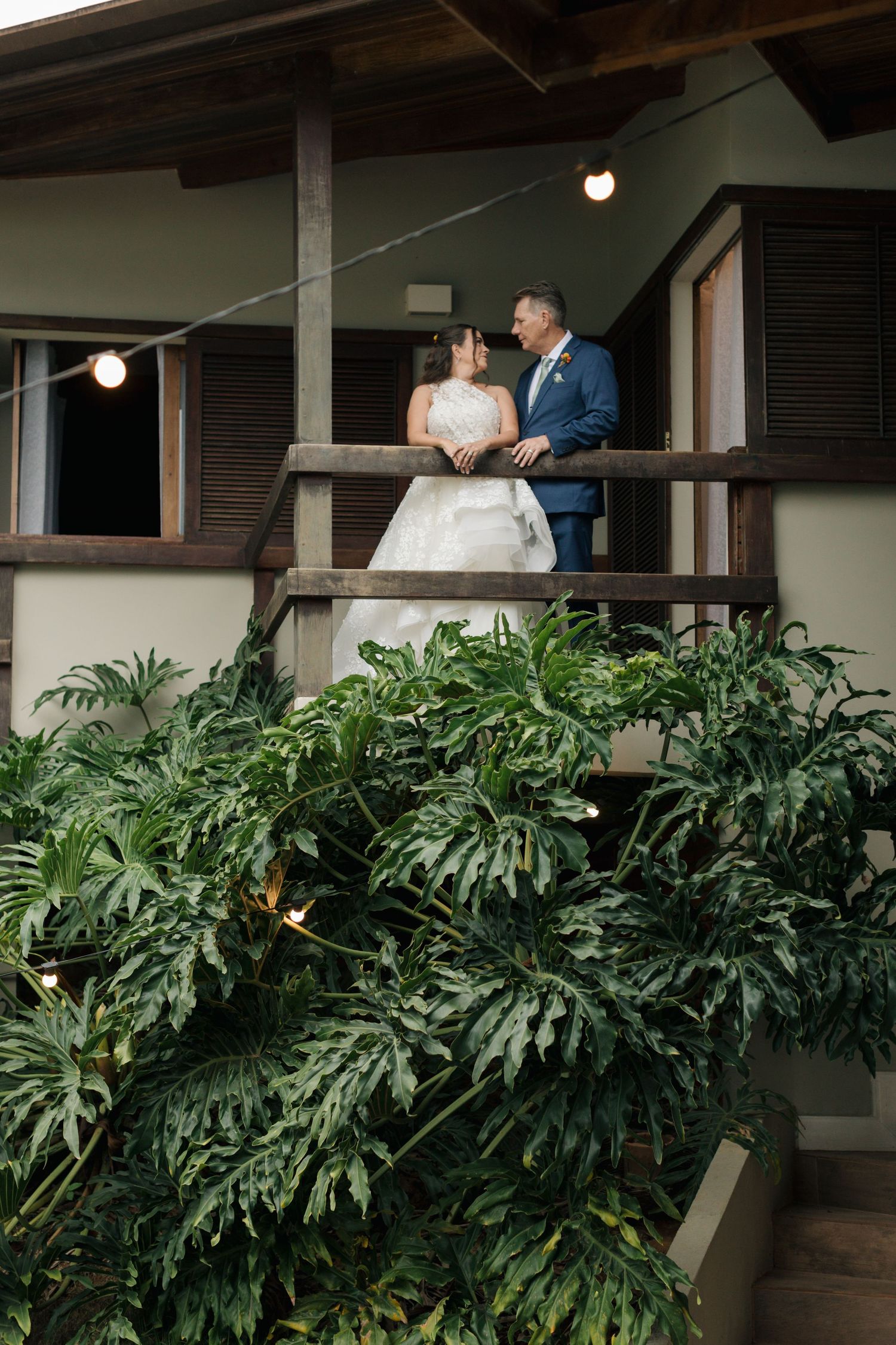 A romantic wedding couple embraces on a covered porch surrounded by lush tropical monstera plants and greenery.