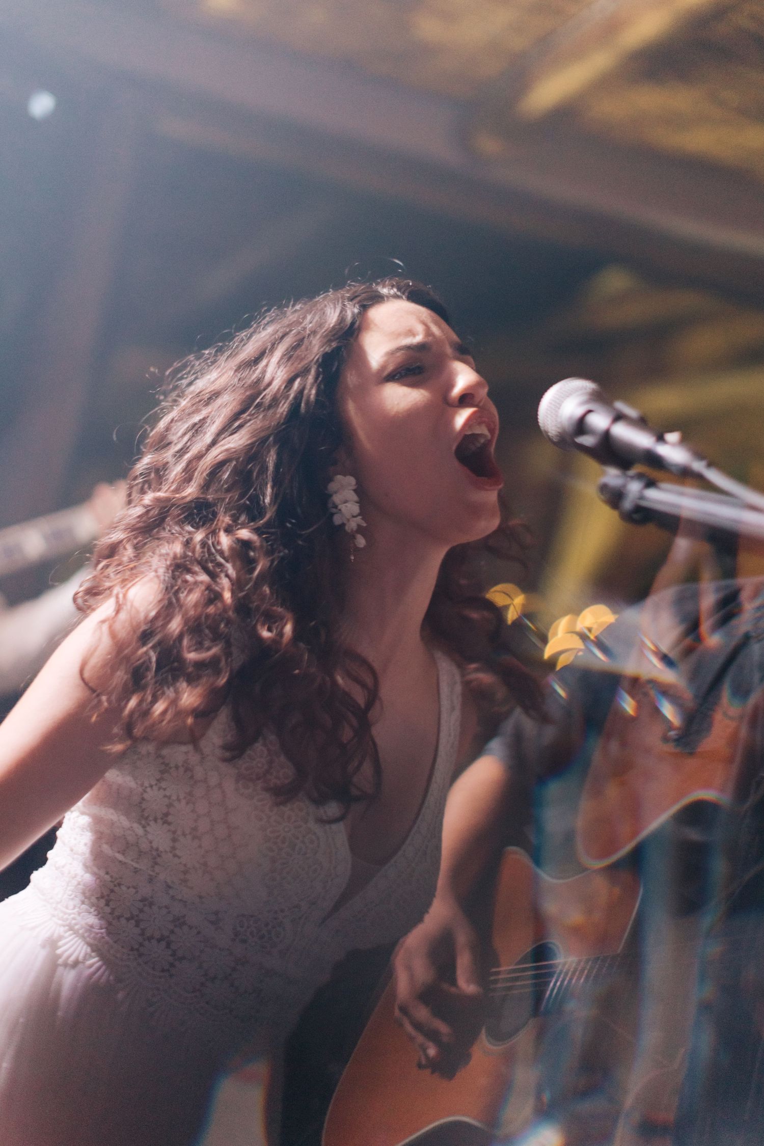 Performer in white dress sings emotionally into microphone during live musical performance.