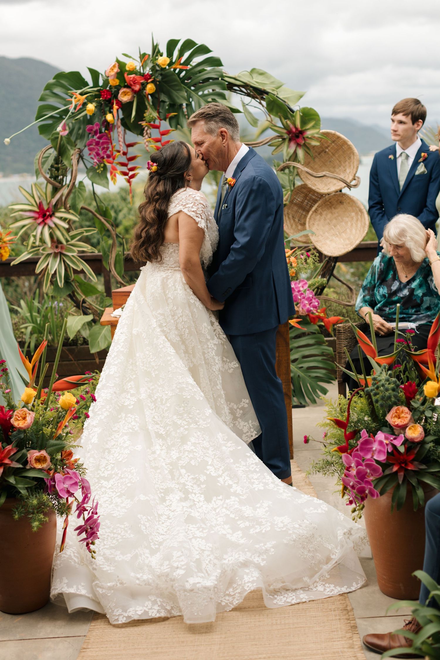 A tropical wedding ceremony setup with colorful flowers, palm leaves, and woven decor against a mountain backdrop.