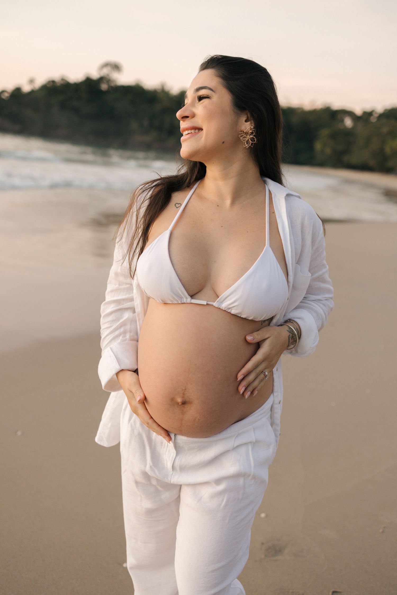 Expectant mother in white beachwear smiling while cradling her baby bump on a sandy shore.
