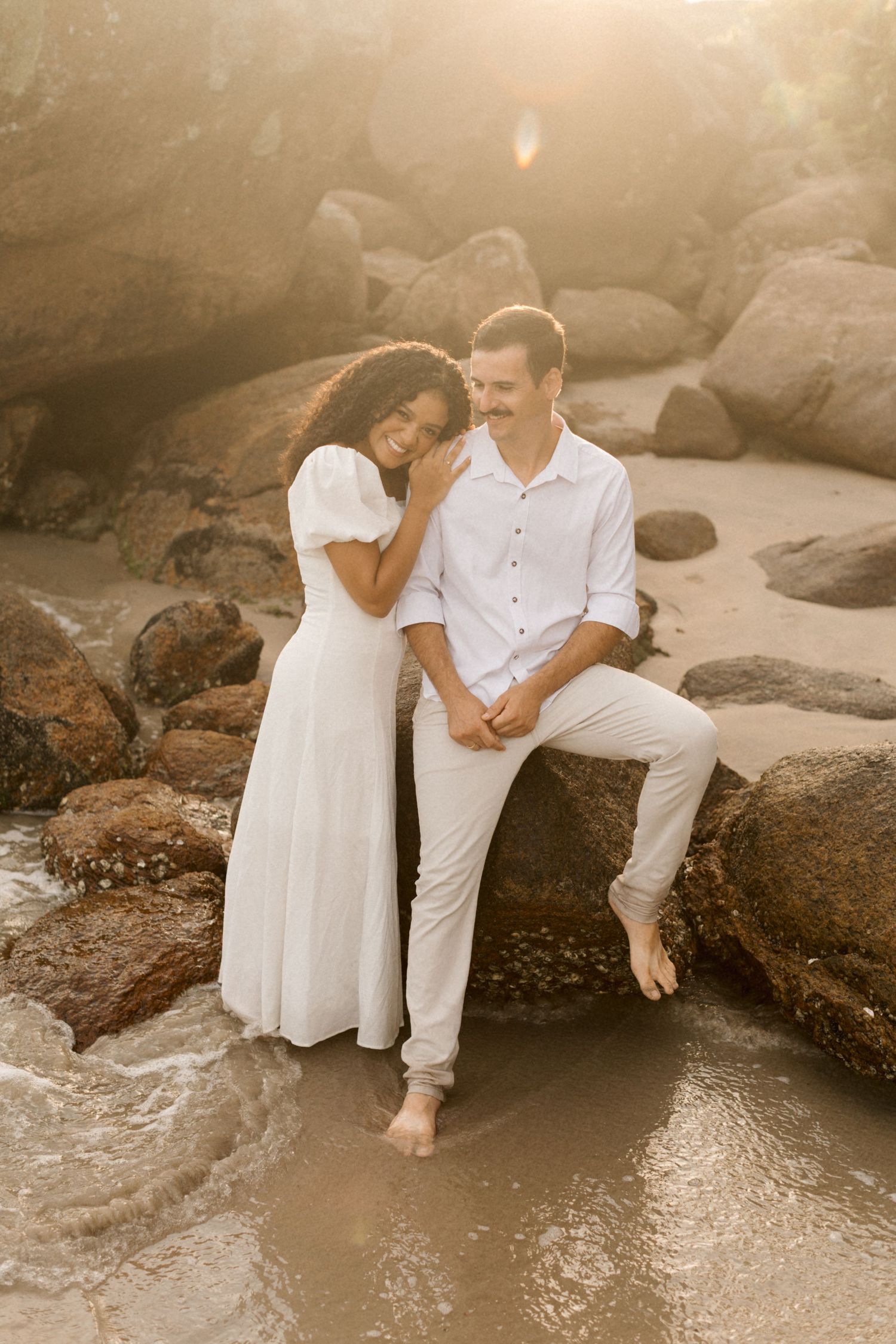 A couple in white outfits sits intimately on coastal rocks near the ocean shore during a photo session.