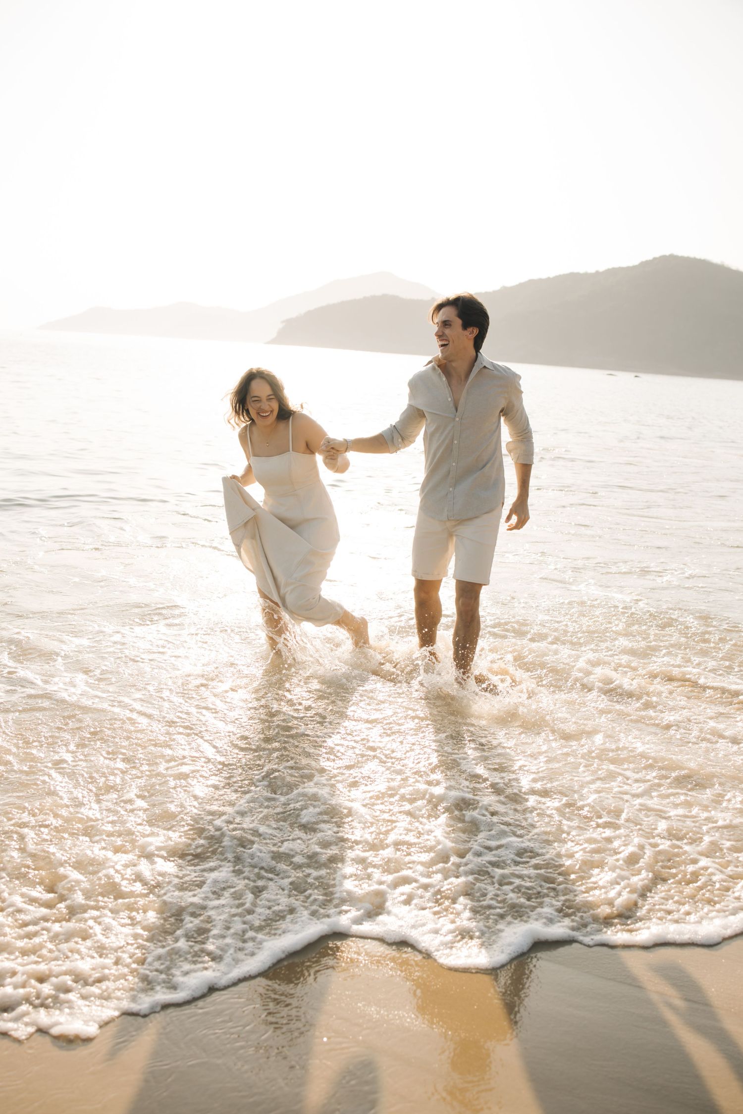 Couple plays in gentle ocean waves at sunset wearing white outfits on a beautiful beach.