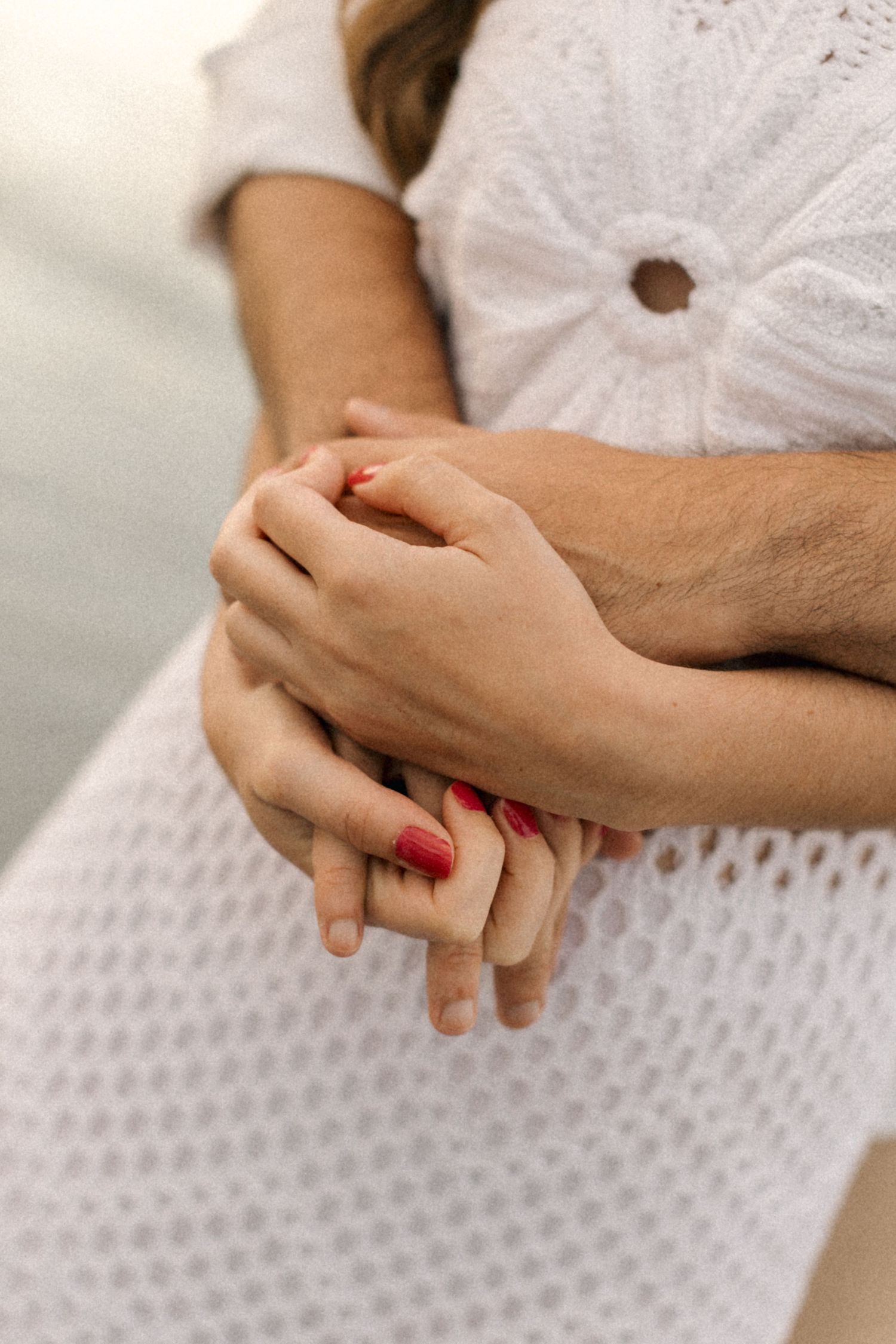 Close-up of intertwined hands against white lace dress showing red nail polish.